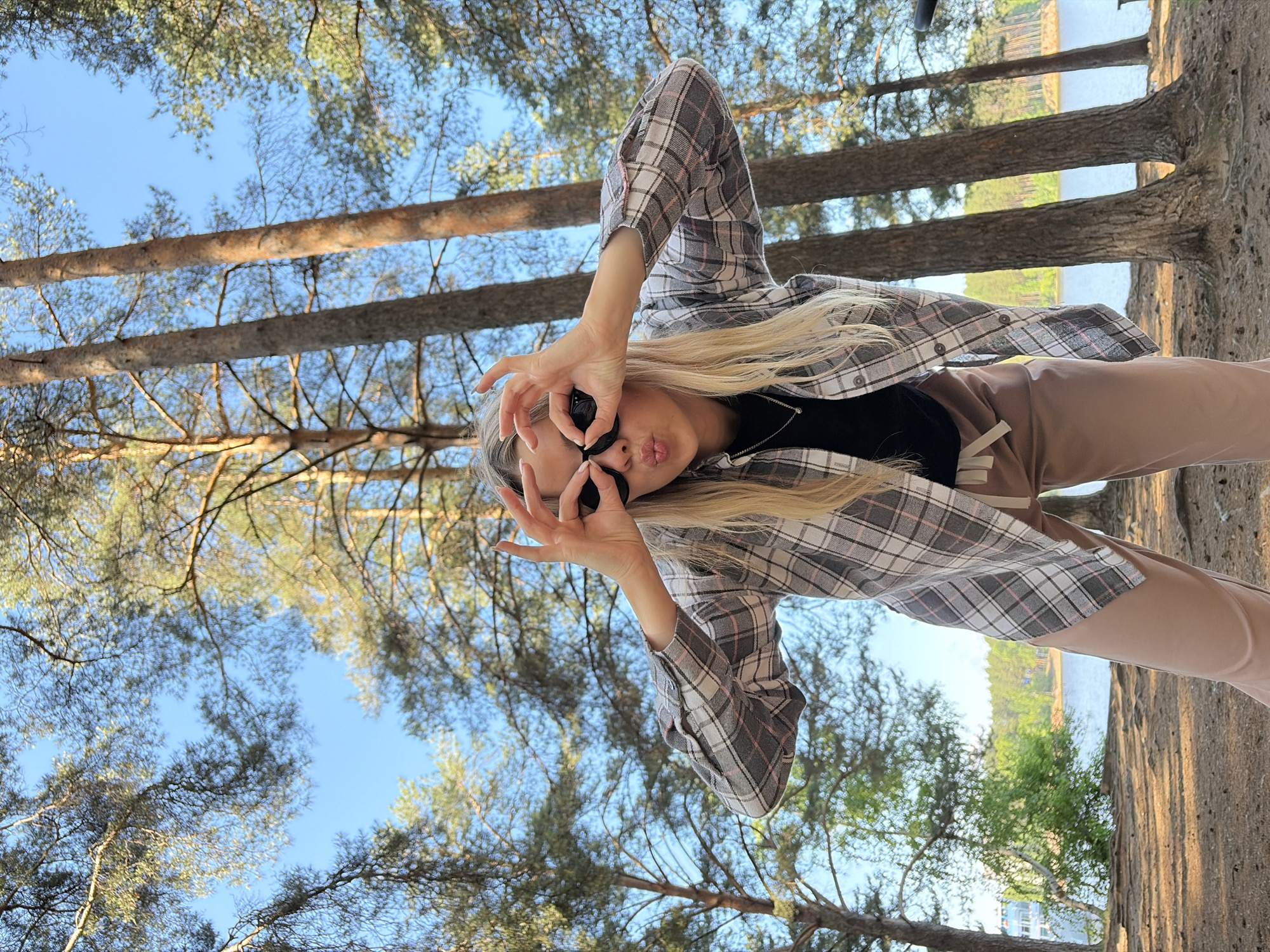 A young woman with blonde hair, wearing a plaid shirt and light-colored pants, is standing outdoors in a forest. She is holding her hands up to her eyes like binoculars and puckering her lips. The background shows tall pine trees, a body of water, and a clear blue sky. The ground is covered in dirt and pine needles. The scene appears to be a natural park or wooded area. This location is consistent with areas near the Kama River in Chaikovsky, Russia, known for its natural beauty and recreational opportunities.