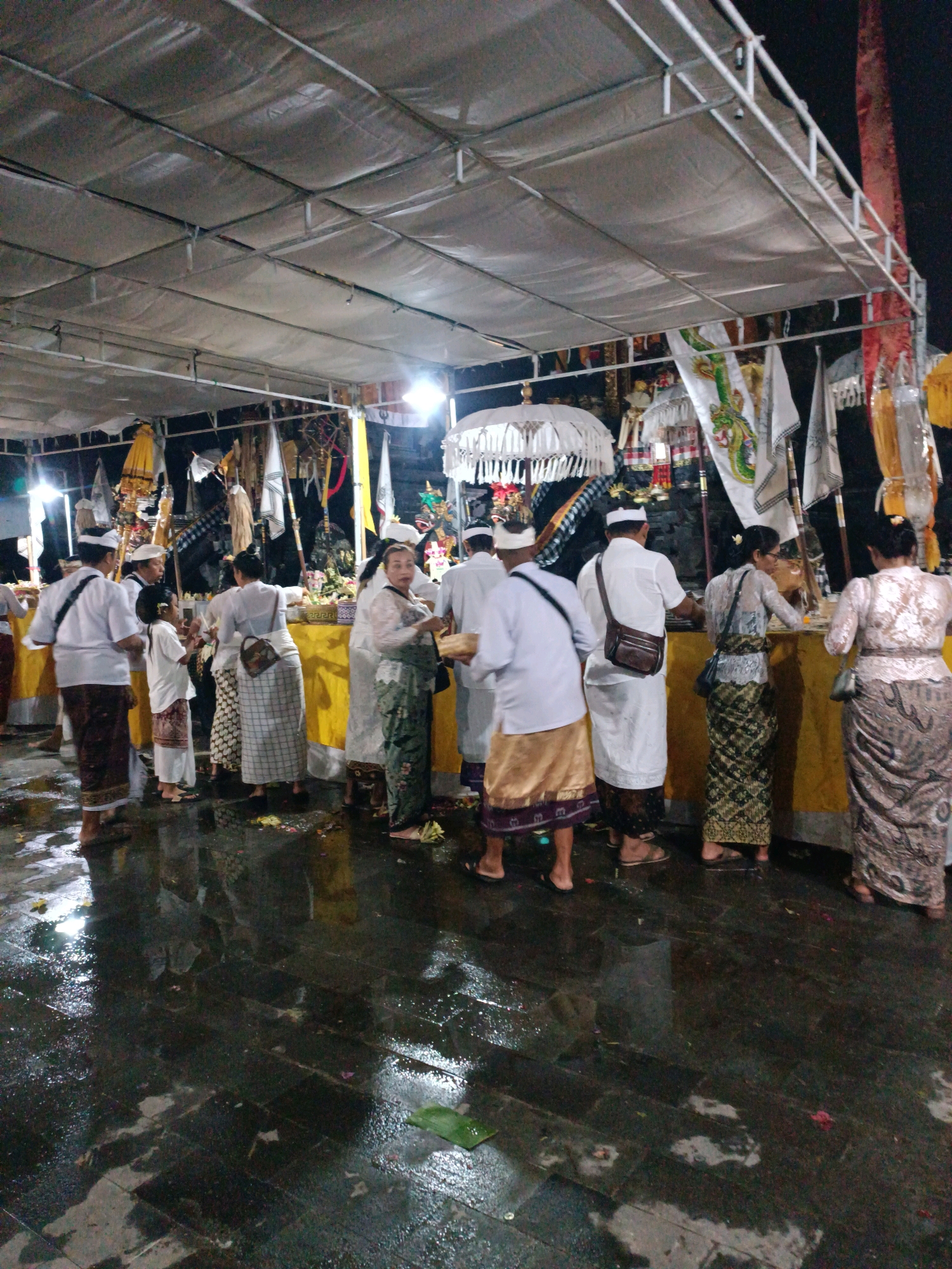 The image depicts an outdoor scene at night, likely during a religious ceremony or event in Kediri, Indonesia. A temporary canopy, supported by metal structures, covers the area. Underneath the canopy, a group of people are gathered, some facing a structure adorned with offerings and flags. The ground is wet and reflective, indicating recent rain or moisture.
Several individuals are dressed in traditional attire, including sarongs and white tops. Some are holding objects or engaging in conversations. In the background, there are decorative elements such as white umbrellas, banners with intricate designs, and possibly religious statues. The lighting appears to be artificial, casting a warm glow on the scene. The overall atmosphere suggests a communal gathering with a spiritual or cultural significance.