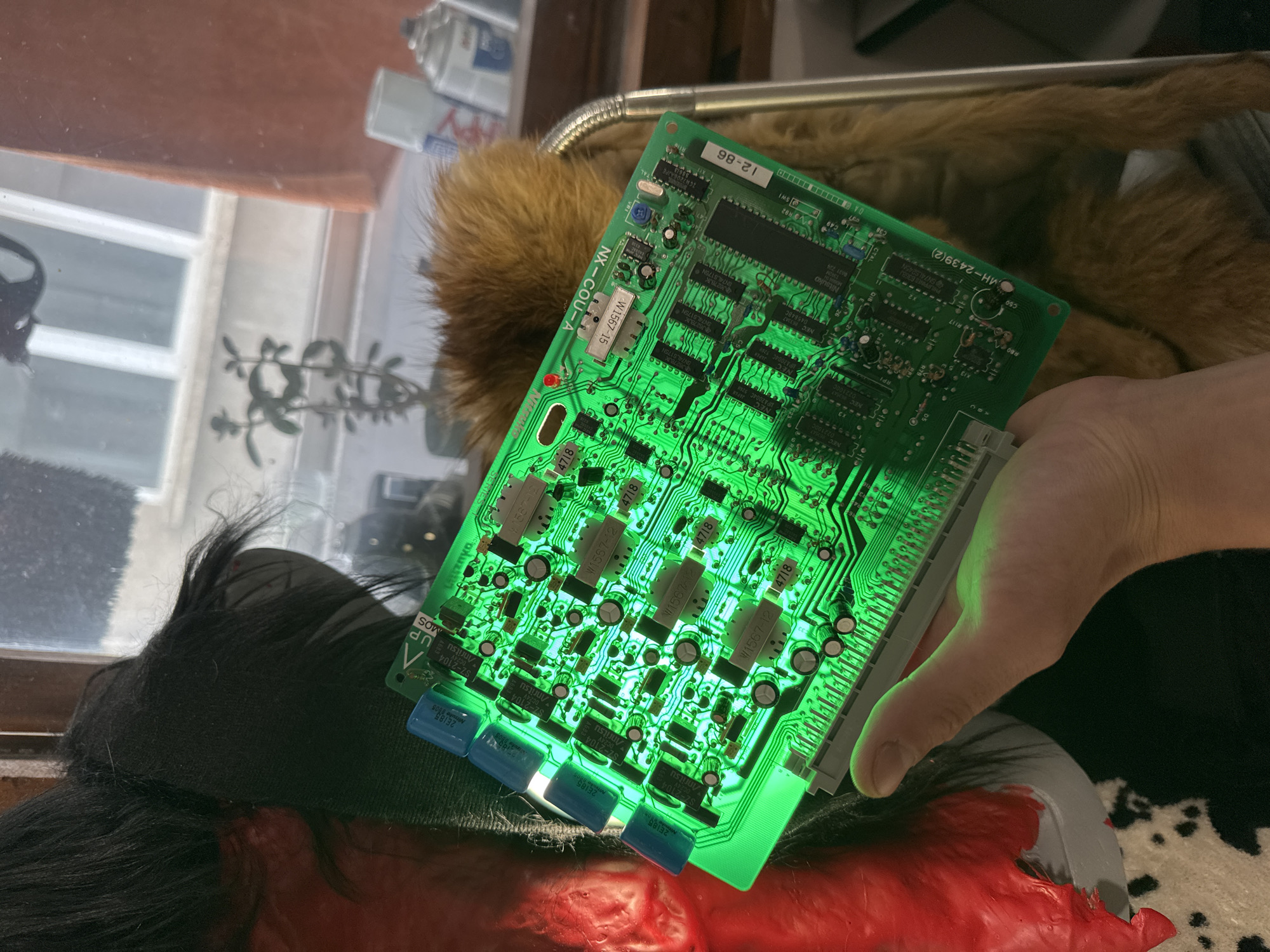 An individual's hand holds a brightly lit green circuit board, exhibiting a distinct glow. The circuit board appears to be part of an electronic device, possibly a gaming machine, given the visible components and connectors. In the background, a window shows a glimpse of an urban environment with trees and buildings, hinting at a city setting. The interior of the room appears somewhat cluttered, with various objects and textures visible, including what seems to be a stuffed animal or fur coat, and a shelf with spray cans. The overall impression is of a casual, possibly domestic, environment where the circuit board is being examined or worked on. The city of Sofia, Bulgaria, is not directly referenced or depicted in the image, therefore no specific location within Sofia can be identified.