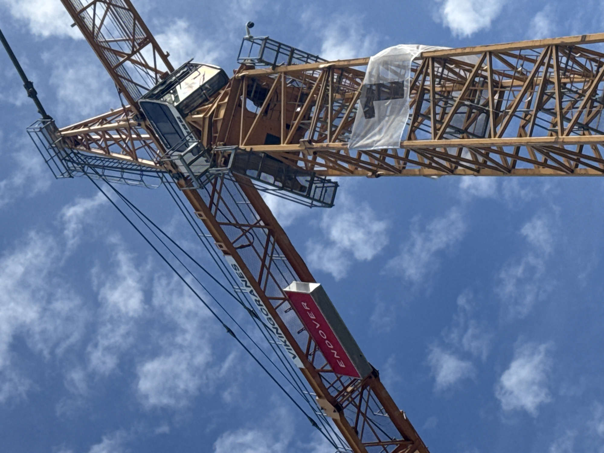 A yellow construction crane stands against a blue sky with white clouds. The crane has a red sign that reads "Endover" hanging from its jib. Further up the crane, on its tower, a white banner with a black number "1" is attached. The crane is positioned in an outdoor environment, likely a construction site.