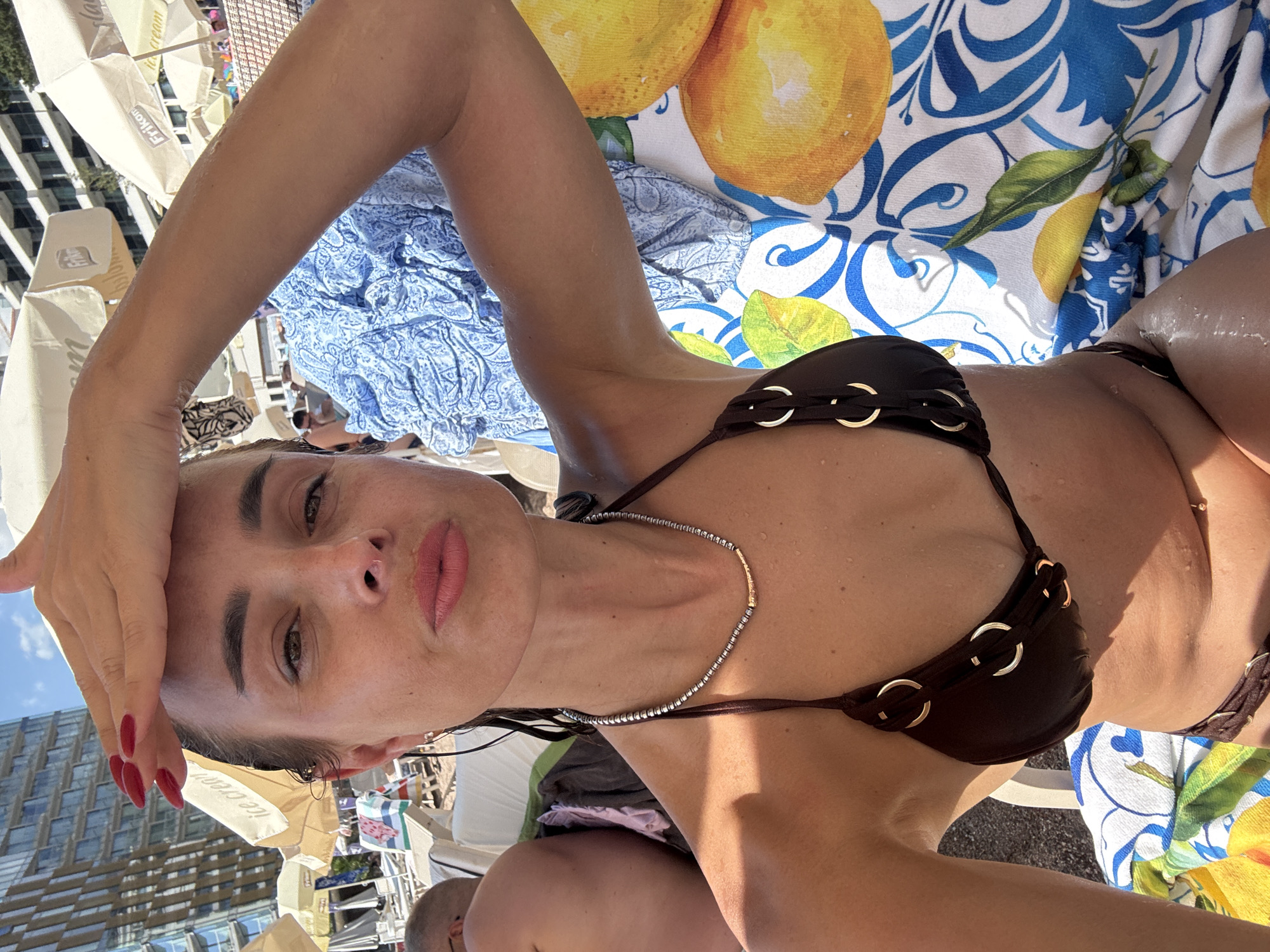 This is a vertical, medium shot of a woman lying on her back, facing the camera. She is outdoors, likely at a beach or pool, as indicated by the presence of lounge chairs, umbrellas, and a patterned towel with lemons. The sun is shining brightly, creating strong shadows and highlights, suggesting it is daytime. The woman is wearing a brown bikini and a delicate necklace. Her right hand is raised and resting on her forehead, and her skin appears slightly dewy, perhaps from sweat or water. Her expression is neutral. In the background, there are glimpses of buildings and more beach umbrellas, hinting at a resort or urban beach setting. A portion of another person's shoulder is visible in the lower right corner. There is visible text on a white umbrella behind the woman's head that reads "Ice Cream."