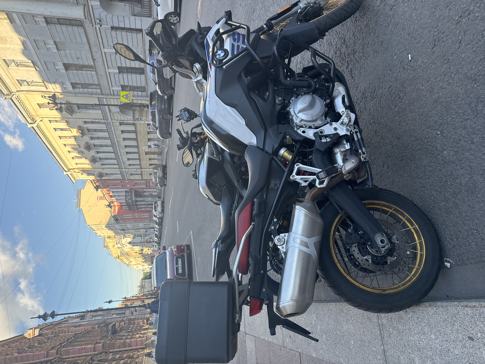 A street scene in Saint Petersburg, Russia, features two motorcycles parked on a sidewalk. The foreground motorcycle is a BMW adventure bike, predominantly silver and black, with a large black storage box mounted on the rear. Its front wheel is visible, featuring a gold rim. Behind it, another motorcycle, partially obscured, appears to be a sportbike with a red seat. The background shows a street lined with historic buildings, some with ornate facades and arched windows, bathed in warm sunlight. Several cars are parked along the street, and a few pedestrians can be seen walking on the sidewalk. The sky above is a clear blue with scattered white clouds.