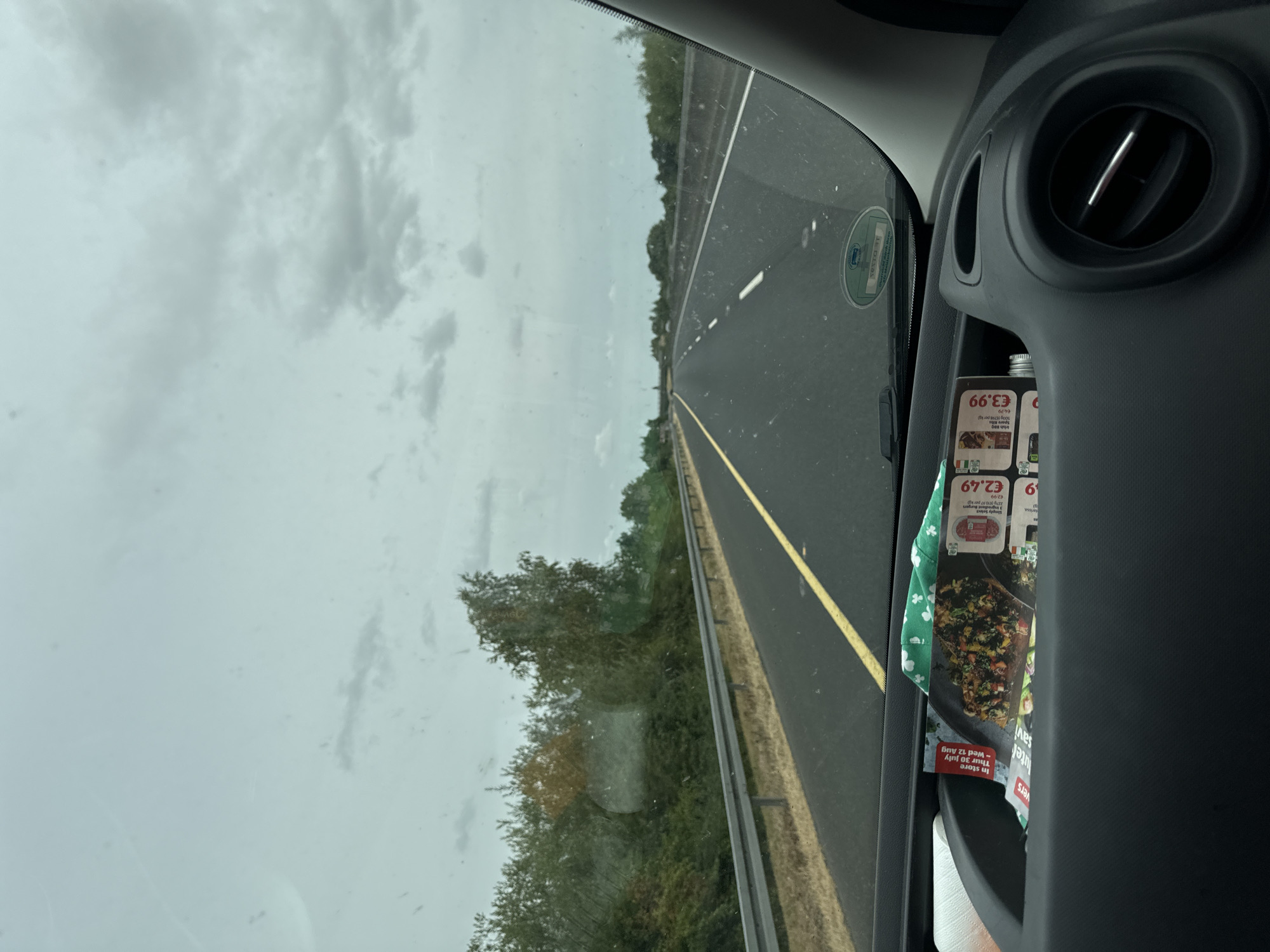 The image is taken from the perspective of a passenger inside a vehicle traveling on a highway. The road ahead is paved asphalt with a yellow line marking the center. On the left side of the road, there is a metal guardrail separating the highway from a grassy embankment covered with trees and bushes. The sky is overcast with clouds. Inside the vehicle, on the dashboard, there is a compartment containing what appears to be promotional flyers or magazines, a roll of paper towels, and a green cloth with a shamrock pattern. One flyer prominently displays an image of a salad and prices such as "€2.49" and "€3.99". A circular green and white sticker is visible on the windshield. The image was captured in Garryhill, Ireland.