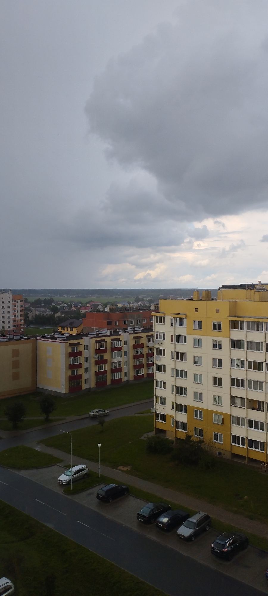 The image depicts an exterior urban scene dominated by overcast skies and apartment buildings. In the foreground, a street runs horizontally with several parked cars. To the left of the street, there are patches of green grass and two streetlights. Further back, across the street, are more grassy areas and a sidewalk.
The middle ground features several multi-story apartment buildings. One prominent building is tall and yellow with white accents and numerous windows. Adjacent to it is a shorter, yellow building with red accents around the windows and balconies. Another building in this area is primarily orange-red.
The background shows a low-rise residential area with smaller houses and more apartment buildings, stretching towards the horizon. Beyond the buildings, there is a dark green line of trees and vegetation, followed by a hazy, lighter green expanse, possibly fields.
The sky is heavily clouded with dark gray clouds, suggesting a potentially stormy or rainy day. The overall lighting is dim and diffused. The specific location is identified as Skidzieĺ, Belarus, based on the provided information, though the image itself does not contain any explicit textual identifiers for this location. The scene appears to be a typical residential block in a Belarusian city.