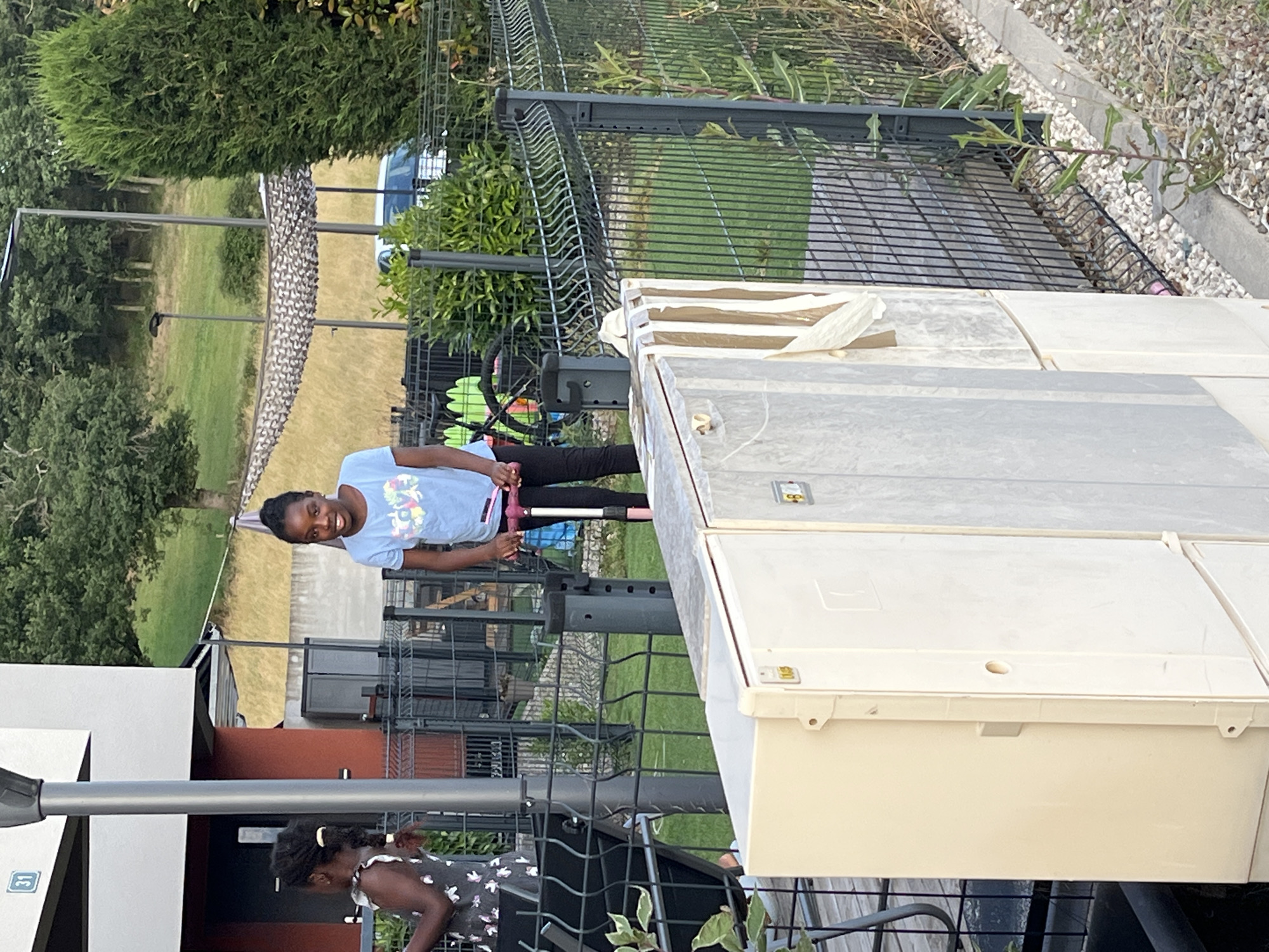 In Chambœuf, France, a young Black girl with braided hair stands on a pink and black scooter, smiling. She wears a light blue t-shirt with a colorful graphic and black pants. To her left, another young Black girl with braided hair in a white dress with a floral pattern is partially visible. They are in a fenced yard with green grass, trees, and a building with a red accent wall and a number "31" visible on a white facade. The background shows a wooded hillside and a white car. In the foreground, large cream and gray utility boxes are present. The overall scene depicts a residential outdoor setting on a sunny day.