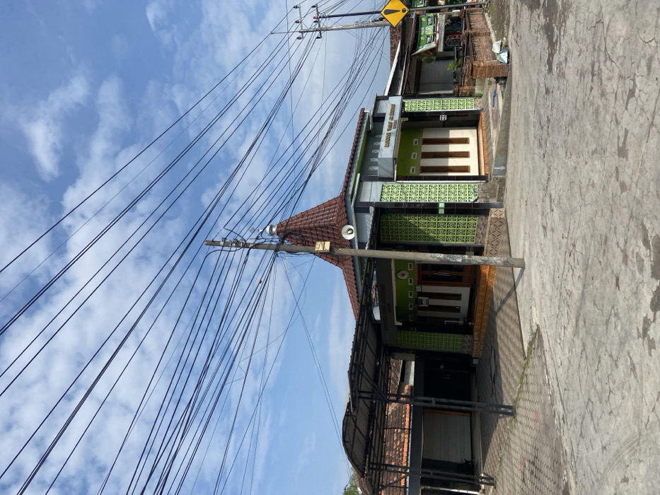 The image shows a mosque located at coordinates -7.498, 112.689 in Kecamatan Tanggulangin, Indonesia. The mosque has a green tiled exterior and a brown tiled roof with a small dome on top. The building is surrounded by a paved area and a small courtyard with a concrete ramp leading up to the entrance. Several power lines run across the sky above the mosque. There is a metal canopy covering an open garage area to the left of the mosque. In the background, there is another building and a power pole with several electrical wires. The sky is blue with white clouds. There are no people in the image.