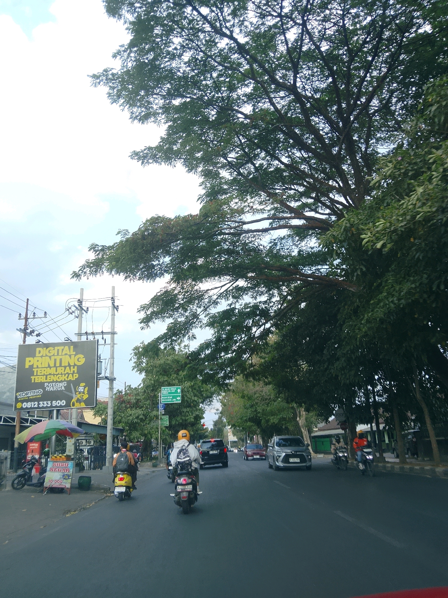 The image depicts a street scene in Kesatrian, Indonesia. A large, leafy tree dominates the upper right portion of the frame, casting shadows onto the road. Several vehicles are visible, including motorcycles and cars. On the left side of the road, there is a prominent billboard advertising "DIGITAL PRINTING TERMURAH TERLENGKAP" with a phone number. A street vendor with an umbrella is also present near the billboard. Traffic appears to be moderate. The sky is overcast.
