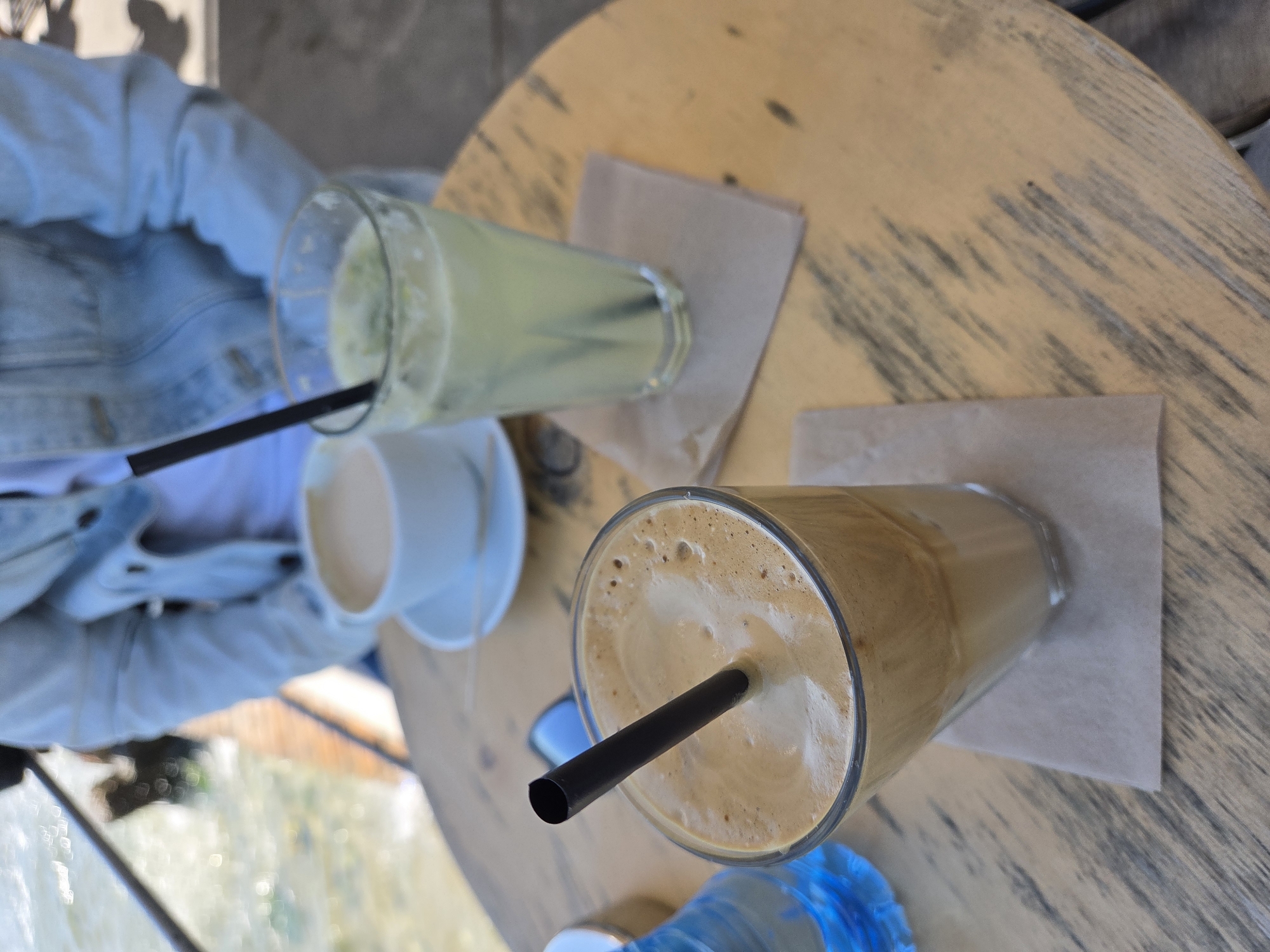 The image presents a high-angle shot of a wooden table, likely in an outdoor café setting. On the table, there are two beverages in glasses: one appears to be a latte or cappuccino with a black straw and foam, placed on a square, tan napkin. The other glass contains a light green liquid, potentially a juice or smoothie, also with a black straw and a tan napkin. To the left of the glasses, there's a white cup on a saucer, possibly containing coffee or tea. A person wearing a light blue denim jacket is partially visible on the left side of the frame. The location is suggested to be in Varna, Bulgaria, but there is no obvious element to confirm this, it can be considered to be on a terrace.