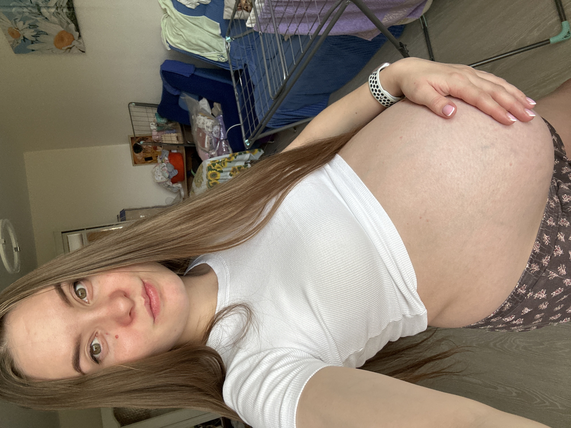 A young woman is taking a selfie in what appears to be a bedroom in Noviy, Russia. She is facing the camera and holding her pregnant belly. She is wearing a white crop top and patterned shorts. The room is cluttered with laundry and furniture.