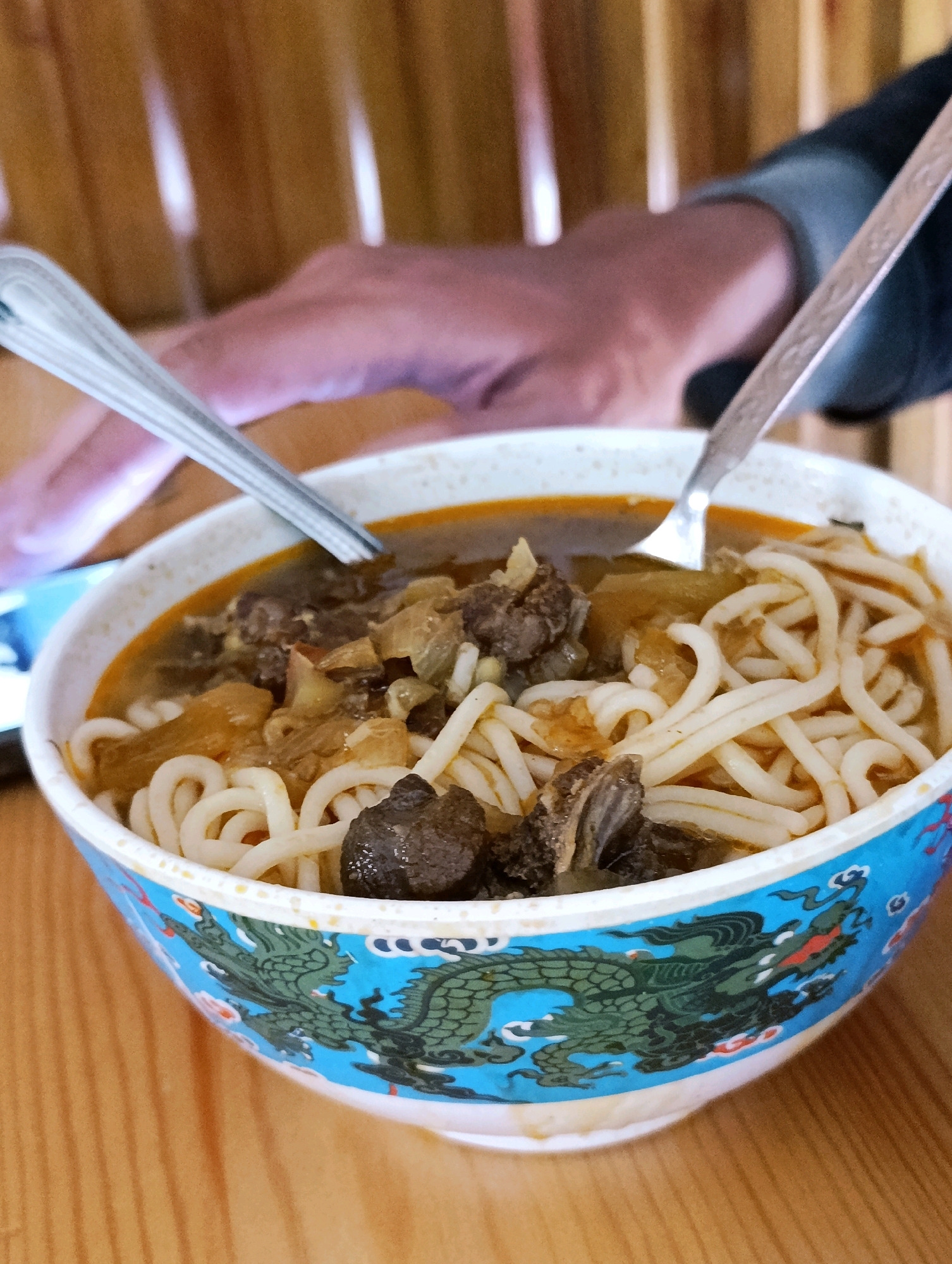A close-up, eye-level shot shows a white ceramic bowl filled with a hearty noodle soup. The soup contains thick, white noodles, chunks of dark meat, and pieces of what appears to be onion or other vegetables in a reddish-brown broth. Two metal spoons are partially submerged in the bowl, suggesting someone is about to eat or has been eating. The bowl is decorated with a vibrant blue band featuring a green dragon motif with red accents. The bowl rests on a wooden surface, likely a table, with a blurred background of vertical wooden slats. The image has a shallow depth of field, with the bowl and its contents in sharp focus. The scene is indoors, possibly a restaurant or home, in the city of Darcha, India.