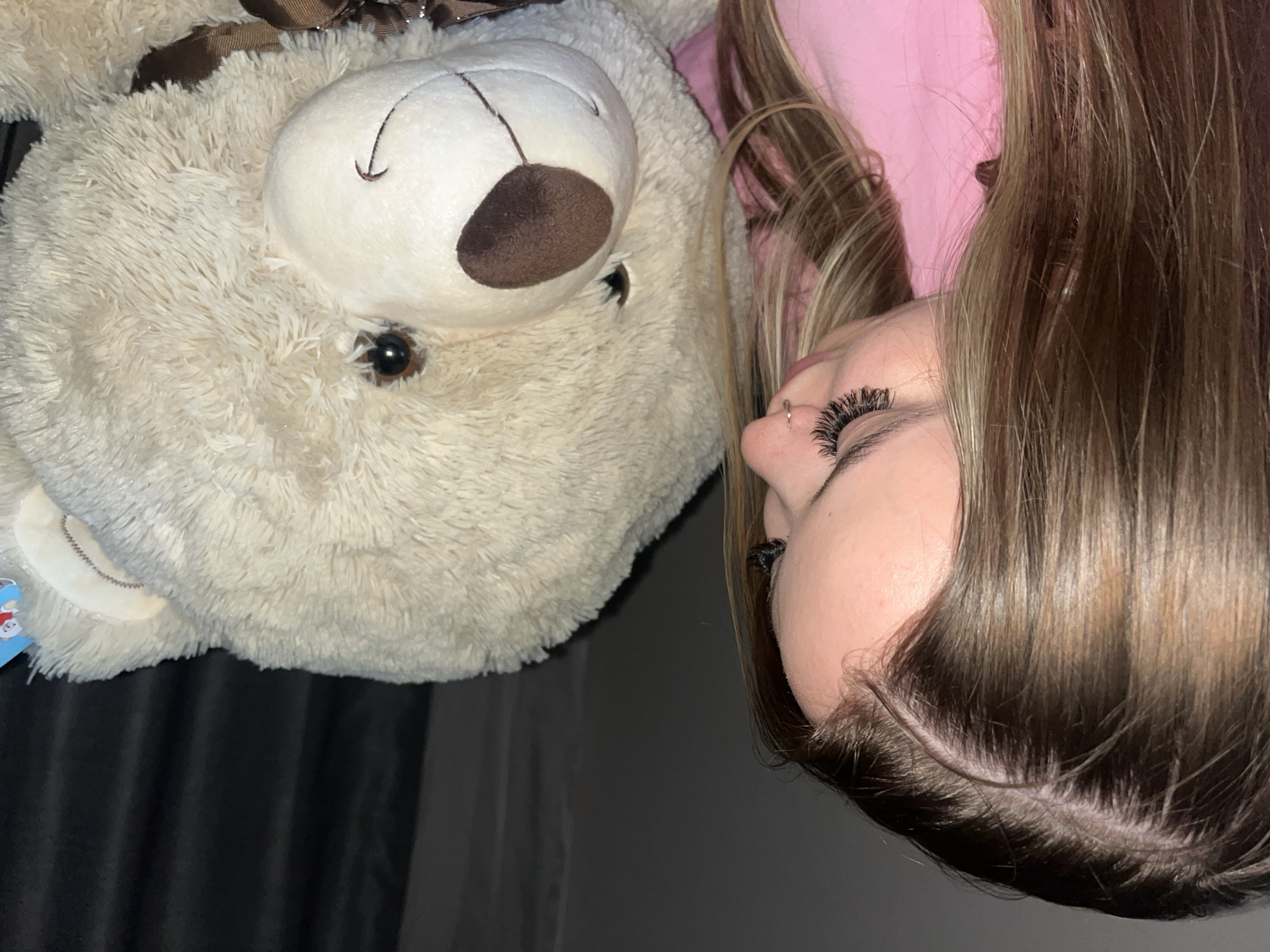 The image presents a close-up, sideways view of a person interacting with a large, fluffy teddy bear. The person, positioned on the right side of the frame, has long, light brown hair with visible lighter highlights, cascading downwards. Their visible features include distinct, long false eyelashes and a small silver ring piercing on their nose. A hint of pink fabric, possibly from clothing or bedding, is visible behind their head.
The teddy bear, occupying the left side of the frame, is a large, cream-colored stuffed animal with soft, shaggy fur. It has dark, round, button-like eyes, a brown fabric nose, and a stitched facial feature on its snout. A brown ribbon, possibly with a subtle patterned texture, is tied around its head or neck. A small blue tag with an indistinct cartoon figure and unreadable text is visible on the bear's left side.
The person's head is tilted towards the bear, creating an intimate and comforting scene, suggesting affection or a moment of rest. The lighting appears subdued, indicating an indoor environment, possibly in the evening. The background behind the teddy bear is dark and uniform, likely a curtain or wall. No clearly legible text is visible in the image apart from the blurred text on the bear's tag. The location context for this image is Krasnodar, Russia.