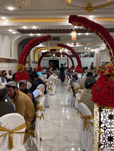 The image depicts a wedding or celebration taking place in what appears to be a banquet hall in Kandahar, Afghanistan. Men are seated at round tables covered with white tablecloths. The chairs are also white, adorned with gold ribbons. Red flower arches with lanterns hang over the tables, adding to the festive atmosphere. Most of the men are wearing traditional Afghan clothing, including shalwar kameez and pakols (woolen caps). The walls are light-colored, and the ceiling has ornate designs. The floor is tiled.