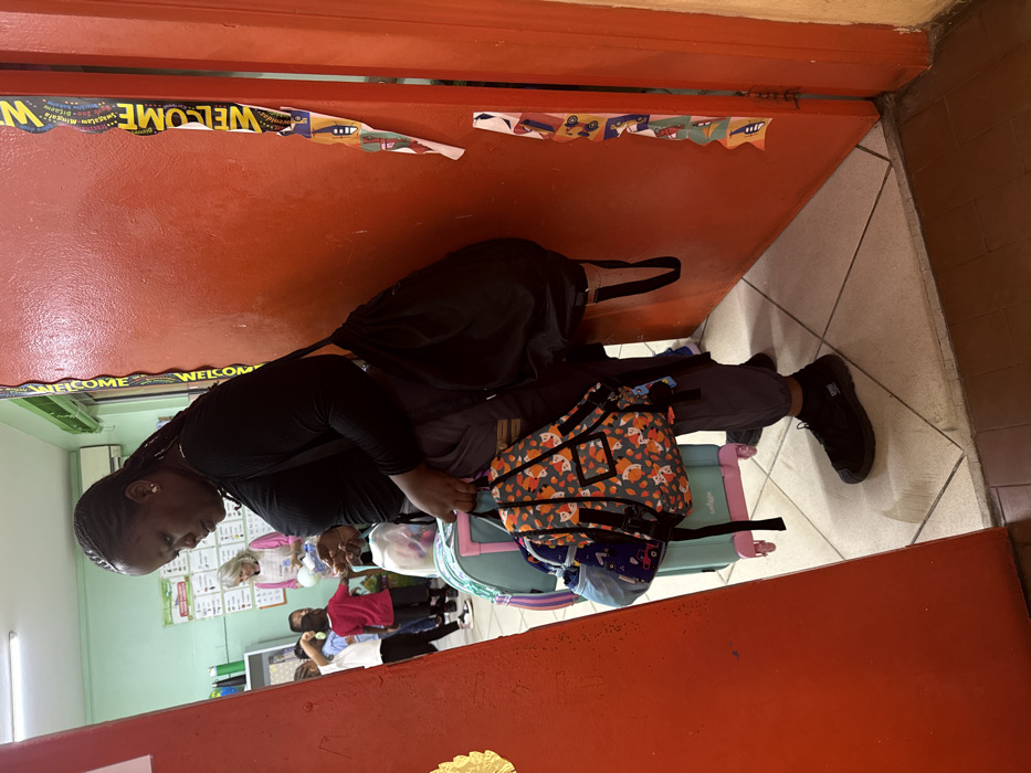 A woman in black stands beside a bright orange doorway adorned with a "Welcome" banner. She has braided hair and wears dark pants and shoes. Beside her are bags, including one with an orange fox pattern and a blue suitcase. In the background, a classroom setting shows students, desks, and wall decor, including a green board. The room is filled with activity and appears to be a vibrant learning environment.