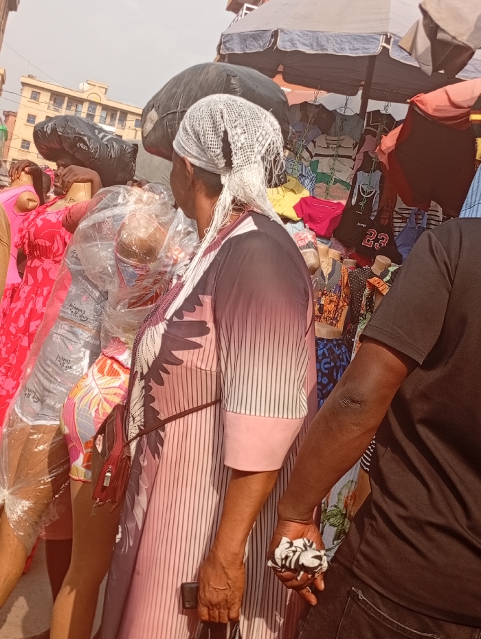 The scene depicts a busy outdoor market in Fegge, Nigeria, under bright daylight conditions. In the foreground, a woman is seen from the back, slightly to the left, wearing a long-sleeved, collared dress with a pink and white striped lower half and a dark, abstract pattern on the upper torso. She wears a white, loosely woven head covering with visible loose strands. A red shoulder bag is visible on her left side, and her right hand holds a dark, rectangular object. To her immediate right, a man's right arm and torso are visible; he wears a dark short-sleeved shirt and holds a crumpled black and white patterned cloth in his right hand. To the left of the woman, multiple store mannequins, some wrapped in clear plastic, display colorful clothing including a bright pink dress, patterned shorts, and grey shorts. Large black bags are positioned on top of market stalls or display structures behind these mannequins. In the background, various garments are hung on display under large market umbrellas, some grey, some reddish-brown. A multi-story building with a light-colored facade is visible in the upper left background under a bright, slightly hazy sky.