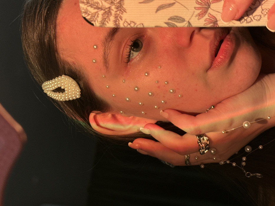 A close-up, angled portrait depicts the left side of an individual's face, likely a young adult female, partially obscured by a smartphone. The subject's left ear is visible, with a white, teardrop-shaped hair clip composed of small pearls fastened in the hair above it. Numerous small, round, white pearl-like beads are adhered to the skin of the subject's left cheek and temple. The subject's right hand (viewer's left), adorned with two rings—one ornate with a dark central design and another a textured band—is positioned with fingers gently touching the left cheek. A string of smaller pearl-like beads is draped across the fingers of this hand. The subject's left eye (viewer's right) is partially visible, featuring a green or hazel iris. The lower part of the nose and the right side of the subject's face are obscured by a vertically held smartphone, the case of which displays a light-colored floral pattern. Strong, warm, reddish-orange artificial light illuminates the visible skin and hair, with a subtle green light perceptible on the far left. The background is dark, indistinct, and out of focus, indicating an indoor setting. The specific location within Krasnodar, Russia, cannot be determined from the visual evidence.