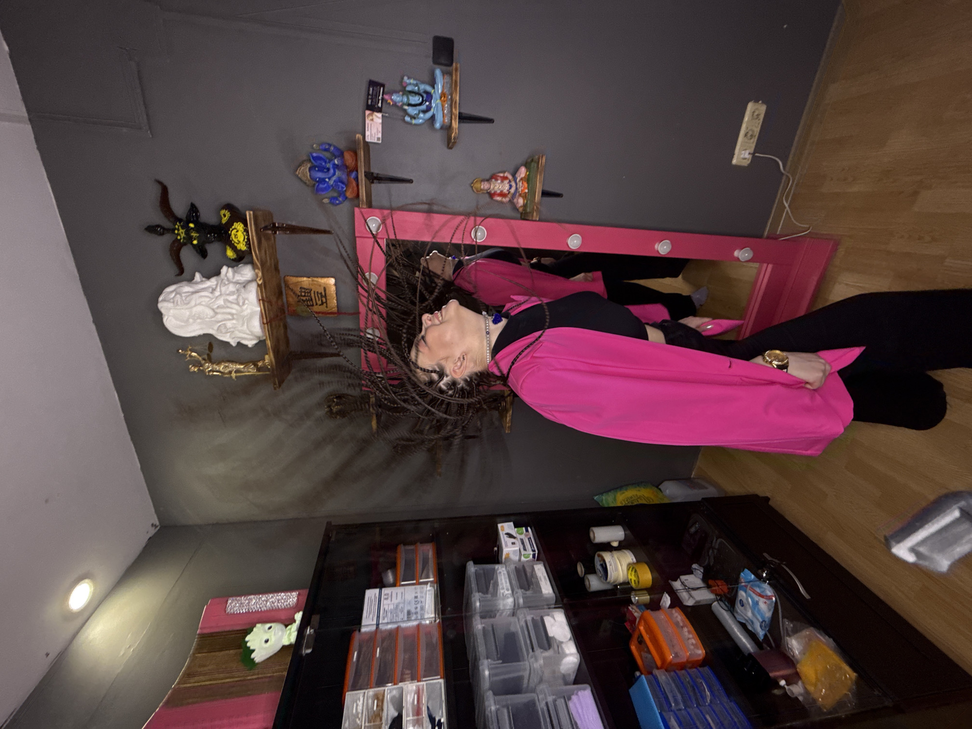 The image depicts an indoor scene in Saratov, Russia. A female subject is centrally positioned, in motion with her head tilted sharply to her left and upwards, causing her long, dark braided hair to fan out and blur around her head. She wears a black sleeveless crop top, black wide-leg trousers, an unbuttoned fuchsia blazer, a white beaded choker with a blue circular pendant, and a gold-toned wristwatch on her left wrist. Her reflection, showing the same posture and attire, is visible in a tall, fuchsia-framed vanity mirror with embedded circular lights, positioned vertically against the right wall.
The room features dark grey walls and light-toned wooden laminate flooring. A single recessed ceiling light illuminates the upper left area. On the right wall, two rustic wooden floating shelves are mounted. The upper shelf displays a golden statue of a female figure holding scales, a white sculpted bust, and a black and yellow horned idol. The lower shelf exhibits three seated figurines: a blue Ganesh, a blue deity figure, and a red deity figure. A golden metallic plaque with an etched symbol is affixed below the upper shelf.
On the left, a dark-stained wood display cabinet with glass doors contains multiple clear plastic storage bins, some with orange lids, filled with various supplies. The top of this cabinet features a light green anthropomorphic figurine and a pink and wood-striped decorative panel. A three-gang electrical outlet with a white cord plugged into one socket is visible on the right wall near the floor.