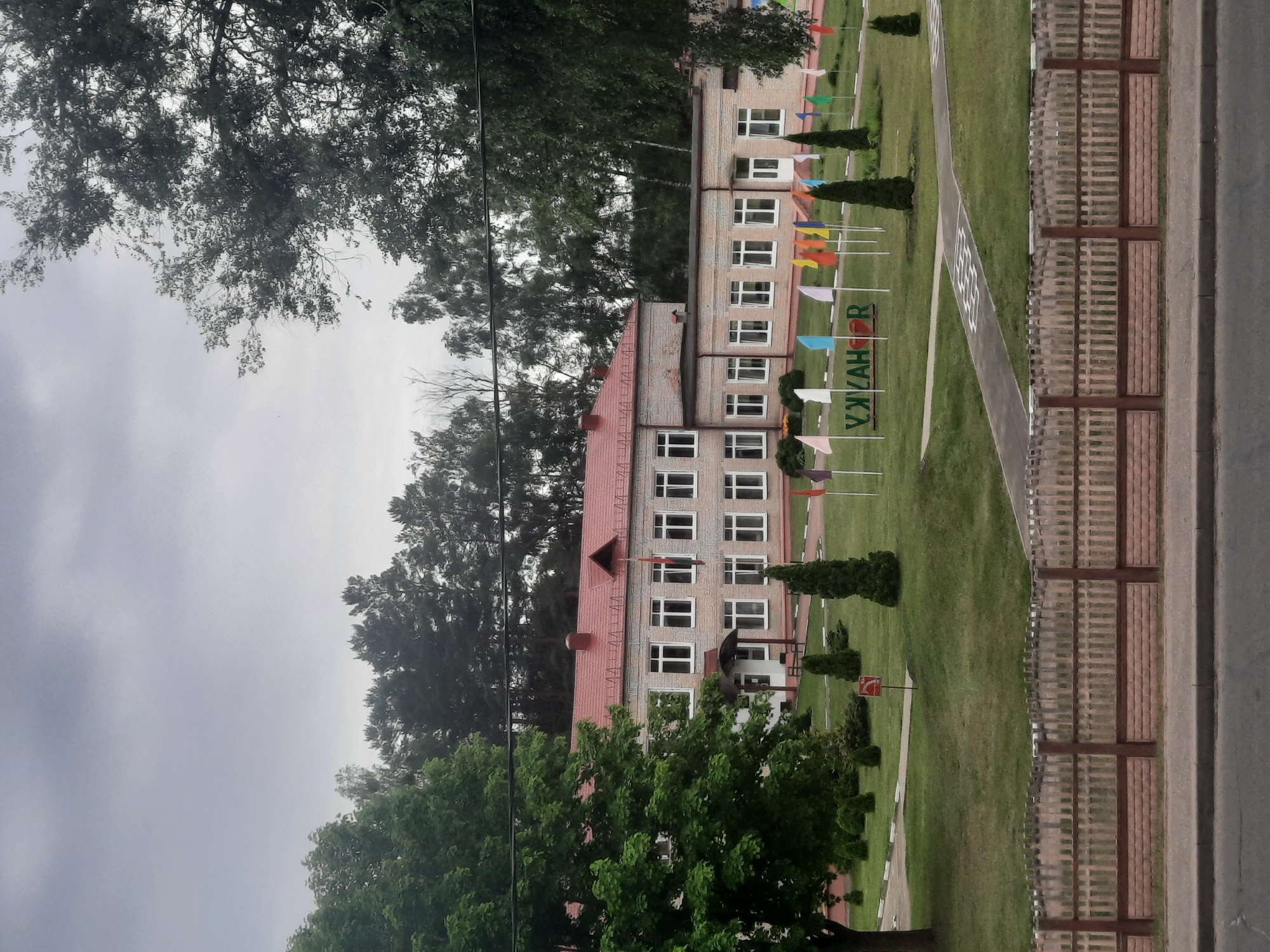 This is a medium-shot, vertically oriented photograph of a school building in Parechcha, Belarus. The building, made of light-colored bricks with a red-tiled roof, has multiple floors and numerous white-framed windows. In front of the school, a neatly mowed green lawn is adorned with small, colorful flags on poles. A paved pathway runs through the lawn, marked with white lines. To the right of the lawn is a wooden fence, and beyond that, a paved area that appears to be a road or sidewalk. Tall, dark green coniferous trees are planted strategically around the school grounds, and the surrounding area is filled with mature deciduous trees with lush green foliage. The sky is overcast and grey, suggesting a cloudy day. There is no visible activity or people in the photograph. The visible text on a green sign reads "УЖАКАР".