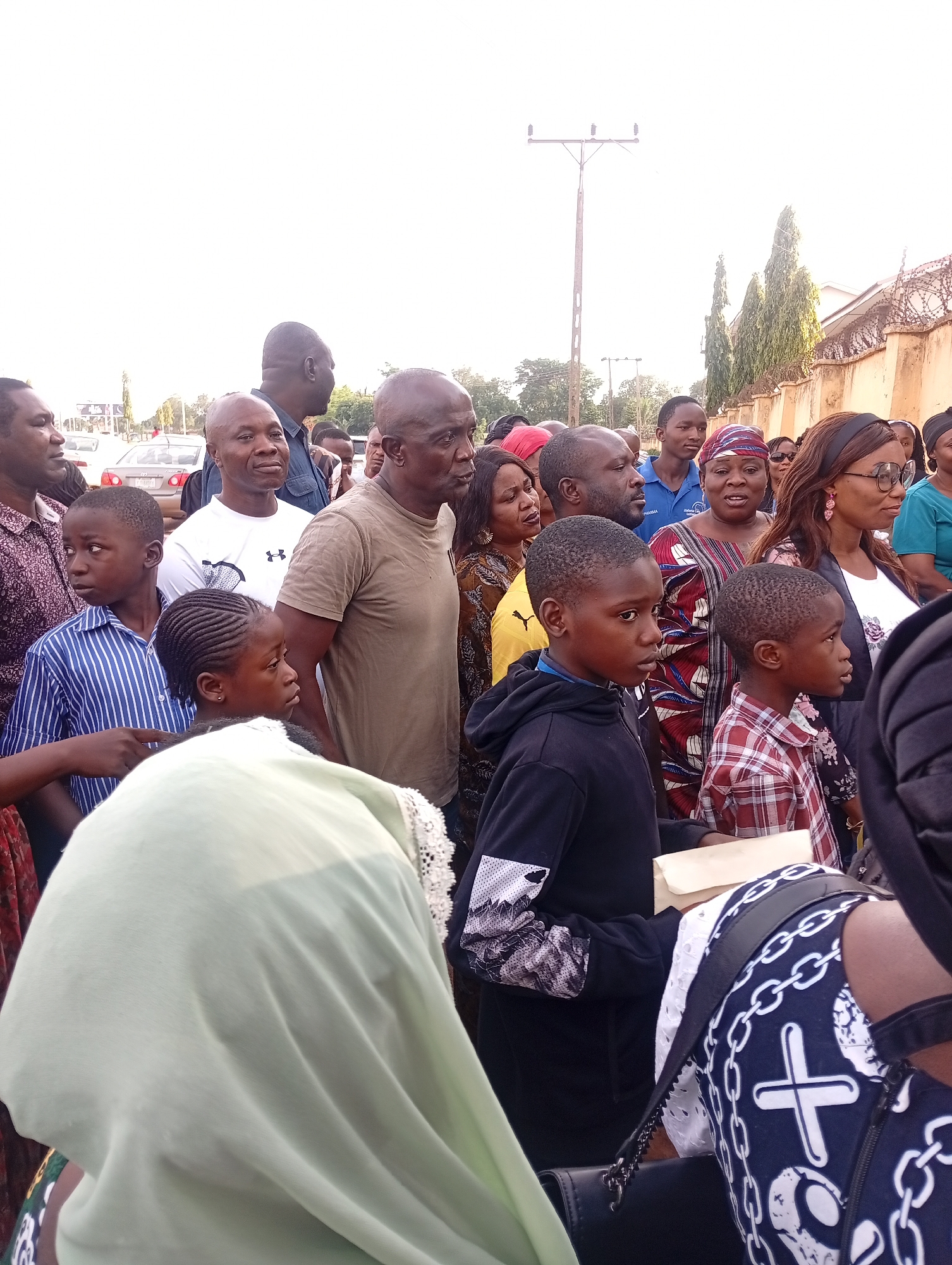 A diverse group of adults and children are assembled in an outdoor setting, possibly in Barden Yana, Nigeria. Most are looking in the same direction. A beige wall with barbed wire is visible on the right. A pole is visible in the background. A vehicle is visible in the distance. People are wearing a variety of clothing.