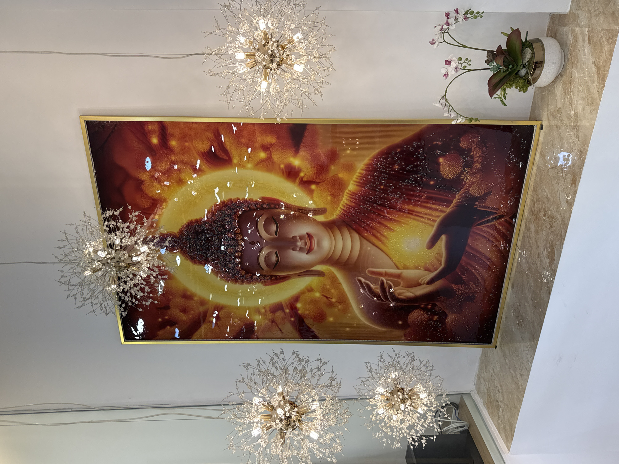 This media file features a ceiling-mounted artwork depicting a serene Buddha. The artwork is framed in gold and displays the Buddha in a lying down position, bathed in a warm, golden light. Multiple sparkling, starburst-shaped chandeliers with crystal accents are visible, suspended from the ceiling, illuminating the scene. To the right of the artwork, a white vase holds artificial orchids and other greenery, placed on a marble-patterned surface, possibly a floor or a counter. The overall setting appears to be an indoor space, possibly a home or a commercial establishment, with clean, white walls and ceiling. The lighting from the chandeliers and the artwork creates a peaceful and elegant ambiance. The time of day is not determinable from the image, and there are no apparent weather cues. The location is identified as Nha Trang, Vietnam. There is no visible text in the image.