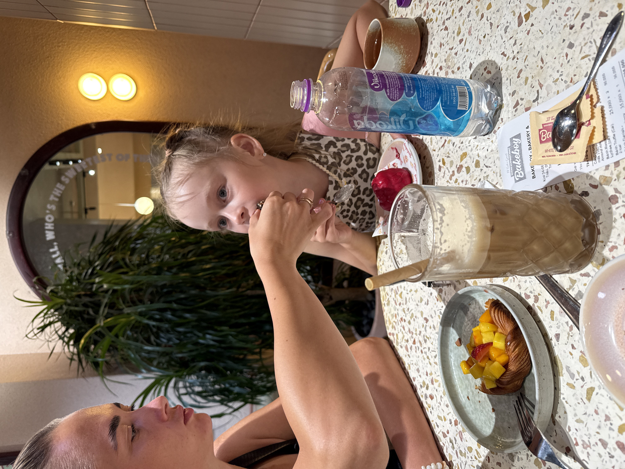 In a brightly lit cafe, possibly located in Nha Trang, Vietnam, a woman is feeding a piece of food to a young girl. The woman, seen from the side, has her arm extended towards the girl, who is looking at the food with her mouth open. The girl has blonde hair in a ponytail and is wearing a leopard-print top. On the table in front of them, there is a pastry topped with fruit, a glass of iced coffee with a bamboo straw, a bottle of water, a small cup of tea, and a receipt from "Bakeboy - Bakery &". The cafe's interior features a large potted plant and a wall with textured tiles. A round mirror with text is visible in the background, and two circular lights are mounted on the wall. The table surface is speckled with various colors.