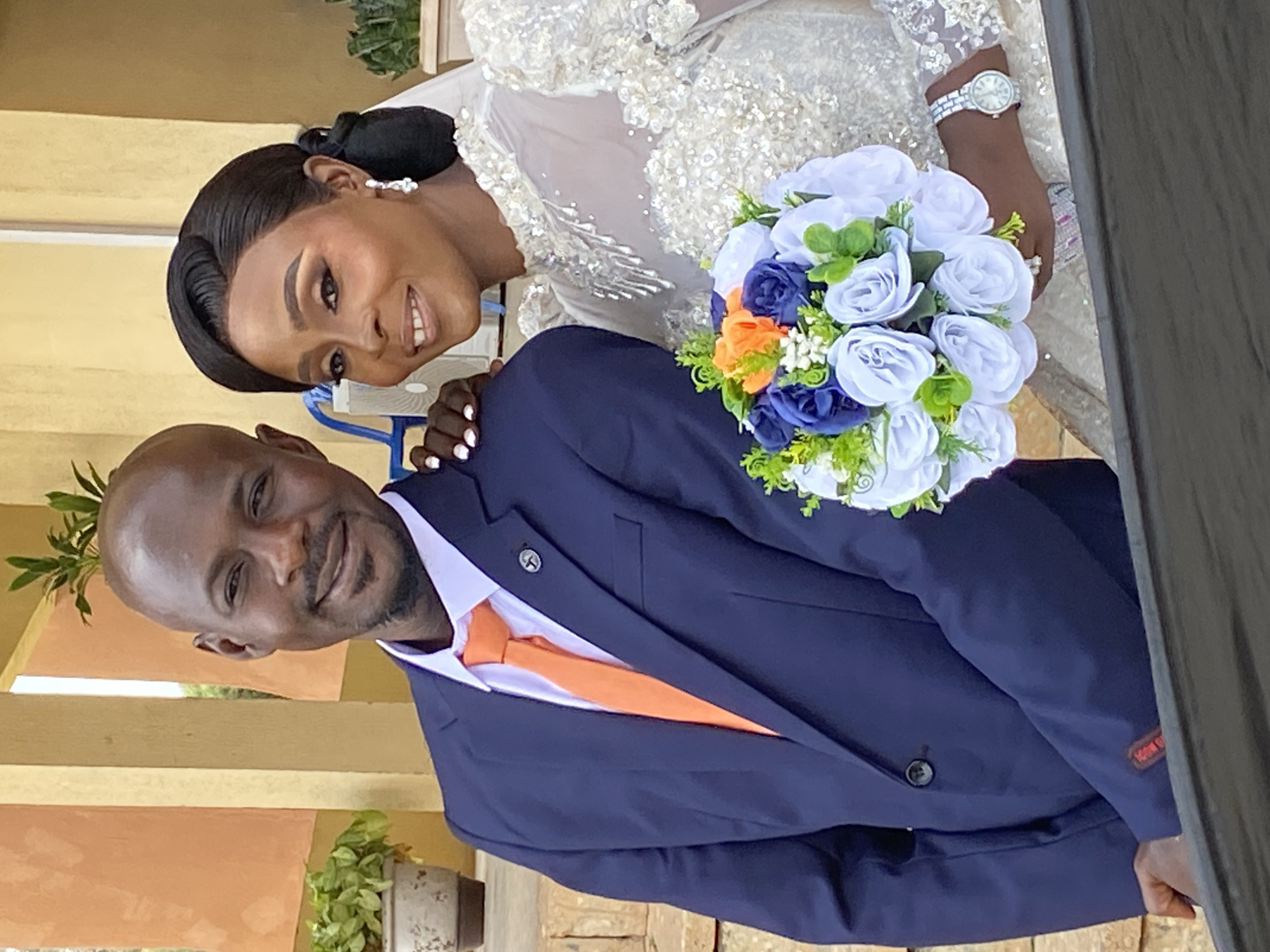 This image captures a wedding portrait of a Nigerian couple, likely taken in Kuru Karama, Nigeria. The bride, adorned in an exquisite white wedding gown with intricate beadwork and lace detailing, leans towards her groom. Her hair is styled in an elegant updo, complemented by pearl earrings. She wears a watch on her left wrist and a ring on her right hand. The groom, dressed in a sharp navy blue suit, white shirt, and an orange tie, smiles warmly at the camera. He has a boutonnière pinned to his lapel. The couple is holding a beautiful bouquet of flowers, featuring shades of white, blue, and orange roses with green foliage.
The background suggests an outdoor or semi-outdoor setting, with hints of yellowish walls and terracotta-colored planters containing greenery. The lighting indicates it's daytime. The overall mood is joyful and celebratory, as evidenced by their smiles and embrace. The image is oriented vertically, with the couple positioned centrally.