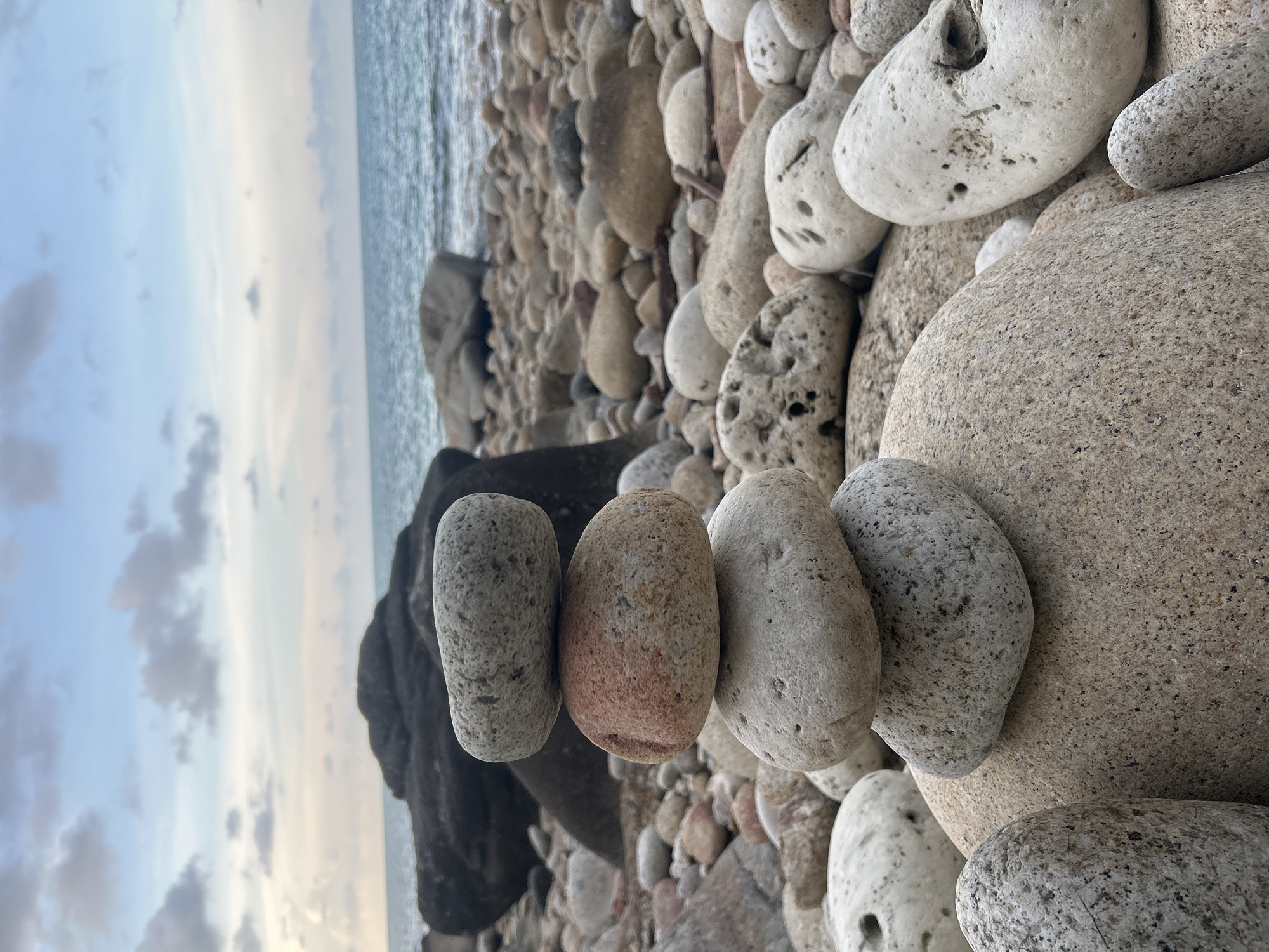 The image depicts a rocky beach in Baan Chaloklum, Thailand. The foreground showcases an arrangement of five smooth, oval-shaped stones, oriented vertically. These stones vary in color, including shades of gray and reddish-brown. They rest atop a bed of similar rocks and pebbles. In the background, the ocean is visible under a cloudy sky. A dark, possibly volcanic rock formation is positioned near the horizon, separating the stones in the foreground from the ocean. There are no people or identifiable subjects other than the rocks and the landscape. The interaction present is the arrangement of the stones. The environment is a natural beach setting at dusk or dawn.