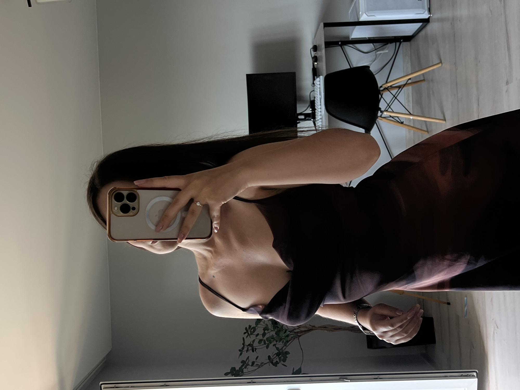 A woman is taking a mirror selfie in an apartment. She is wearing a dark, flowing dress with spaghetti straps. Her hair is long and dark. She is holding a smartphone in her right hand, which is pointed towards the mirror. A ring is visible on her left ring finger. In the background, a desk with a computer monitor, keyboard, and sunglasses is visible. A black chair with wooden legs is in front of the desk. To the left of the woman, a potted plant is situated near a large mirror. The walls are a light gray color, and the flooring appears to be light-colored wood. The overall scene depicts a casual moment within a home environment. It is not possible to determine the specific location within Ternopil, Ukraine.