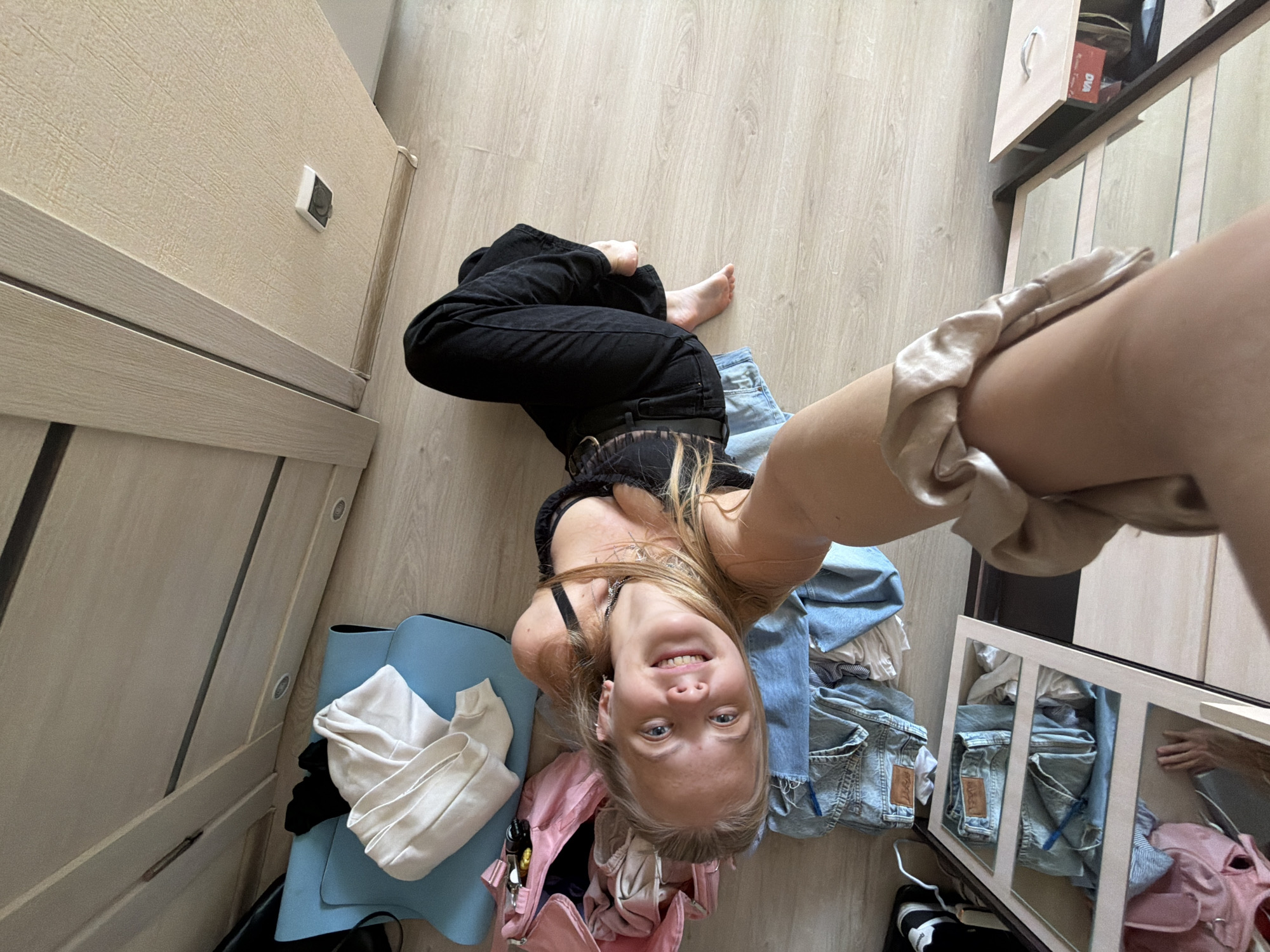 This image, captured in Izhevsk, Russia, is a high-angle selfie taken by a young woman lying on a light wooden floor. Her right arm, holding the camera, and upper thigh are visible in the foreground on the right. The woman has blonde hair, blue eyes, and a wide, radiant smile directed at the camera, conveying a cheerful and relaxed mood. She is wearing a black bra or bralette and black high-waisted jeans, with her bare feet visible.
The scene is an indoor room, likely a bedroom or dressing area, featuring a consistent light wooden laminate floor. Around the woman, various items of clothing are scattered. To her left, a light blue yoga mat or large bag holds a folded white long-sleeved top and some black garments. A pink backpack or bag containing keys and other items is also visible. To her right, a pile of light blue denim clothing, including jeans, is stacked with other fabric items.
The room's furniture includes a light-colored wooden wardrobe or cabinet on the far left, where a wall-mounted light switch or power outlet is visible. On the far right, a tall, light-colored wooden wardrobe with multiple mirrored doors reflects parts of the room, including the denim pile and the pink bag. One of the drawers on this wardrobe is slightly ajar. The overall impression is a casual, personal moment, possibly related to getting dressed or trying on outfits. No clearly legible text is discernible within the image.