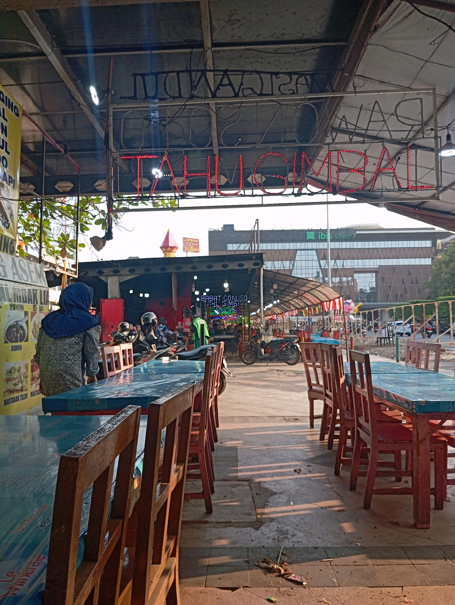 The image depicts an outdoor dining area, likely a warung or food stall, under a covered roof. The scene is set in Semarang, Indonesia, as evidenced by the neon sign above the seating area which reads "Jajanan Tahu Gimbal." The sign uses red neon lights for "Tahu Gimbal" and appears to be older, with a metal frame. Tables covered with blue plastic sheeting and surrounded by wooden chairs are arranged in rows. In the background, there are motorcycles parked, suggesting this is a popular spot. A woman wearing a blue hijab is seated at one of the tables, facing away from the camera. Further in the background, a modern building with the "ibis Styles" logo is visible, indicating its urban setting. The ground is paved with tiles and concrete, with some scattered debris. The lighting suggests it is late afternoon or early evening, with shadows cast by the tables and chairs.