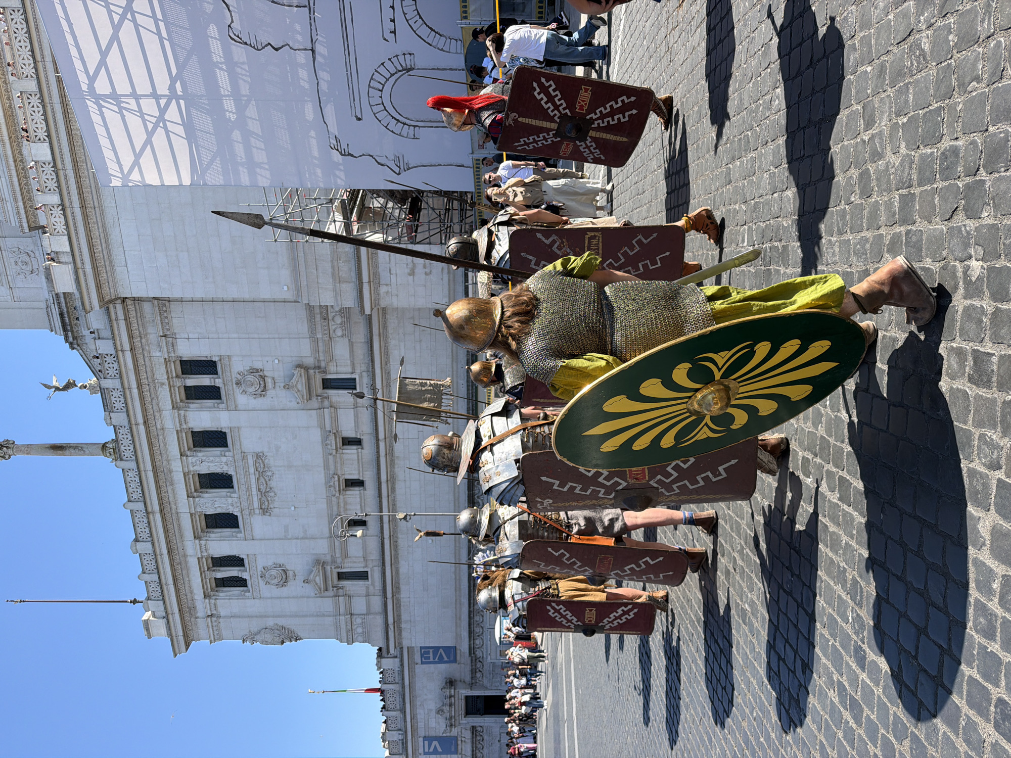 The image shows a line of people dressed in Roman soldier costumes marching on a cobblestone street in Rome, Italy. They are carrying spears and shields and are in front of a large white building with classical architecture. There are other people observing in the background. The sky is blue and clear. Shadows from the people fall on the street.