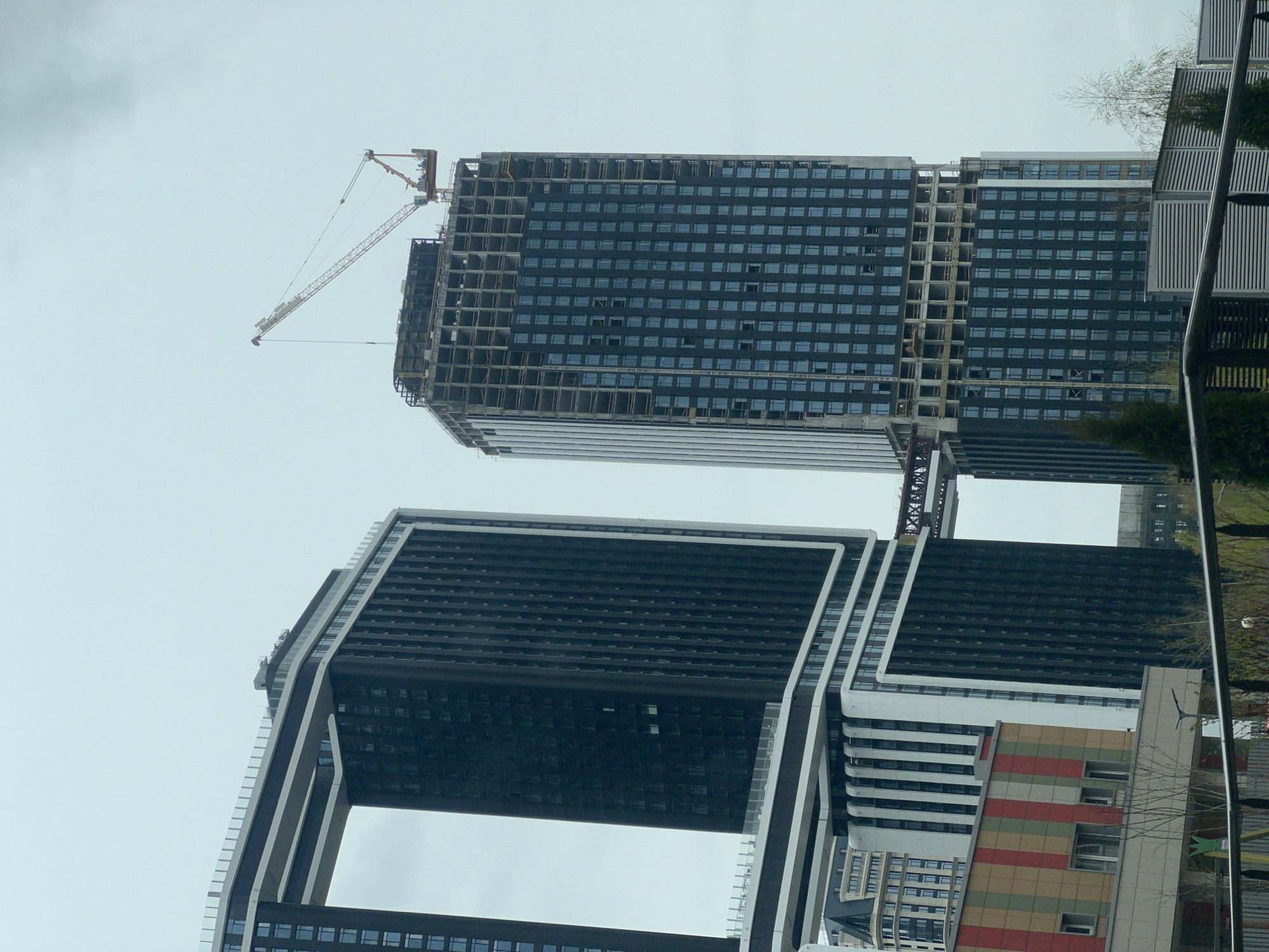 The image shows two tall, modern buildings in Khimki, Russia. One building, topped with a construction crane, appears unfinished. It has a grid-like facade of glass windows. The other building is darker, with a distinctive "U" shape, featuring a large open space in its central section. A neighboring building with colorful horizontal stripes is visible in the foreground. The sky is overcast, and some greenery and architectural details are present along the right edge of the frame.