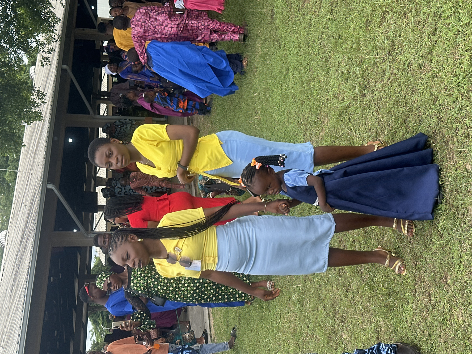 This image depicts an outdoor gathering in Itoga, Nigeria. In the foreground, two adult women and a young girl are the primary subjects. The woman on the left wears a yellow short-sleeved top and a light blue skirt, accessorized with sunglasses on her chest and gold strappy sandals. She holds the hand of the young girl standing to her right, who is dressed in a dark blue dress with puffed sleeves and bows in her braided hair. The woman on the right wears a yellow top and a light blue skirt, similar to the first woman's outfit, and also sports gold strappy sandals. She holds her left hand by her side and her right hand is not visible.
The background reveals a crowd of people dressed in various colorful attire, suggesting a festive occasion. Several individuals are visible under a large, open-sided structure with a corrugated roof, possibly a pavilion or community hall. The ground is covered in green grass, and trees and foliage are visible in the distance, indicating a rural or semi-rural setting. The overall atmosphere appears to be one of community and celebration.