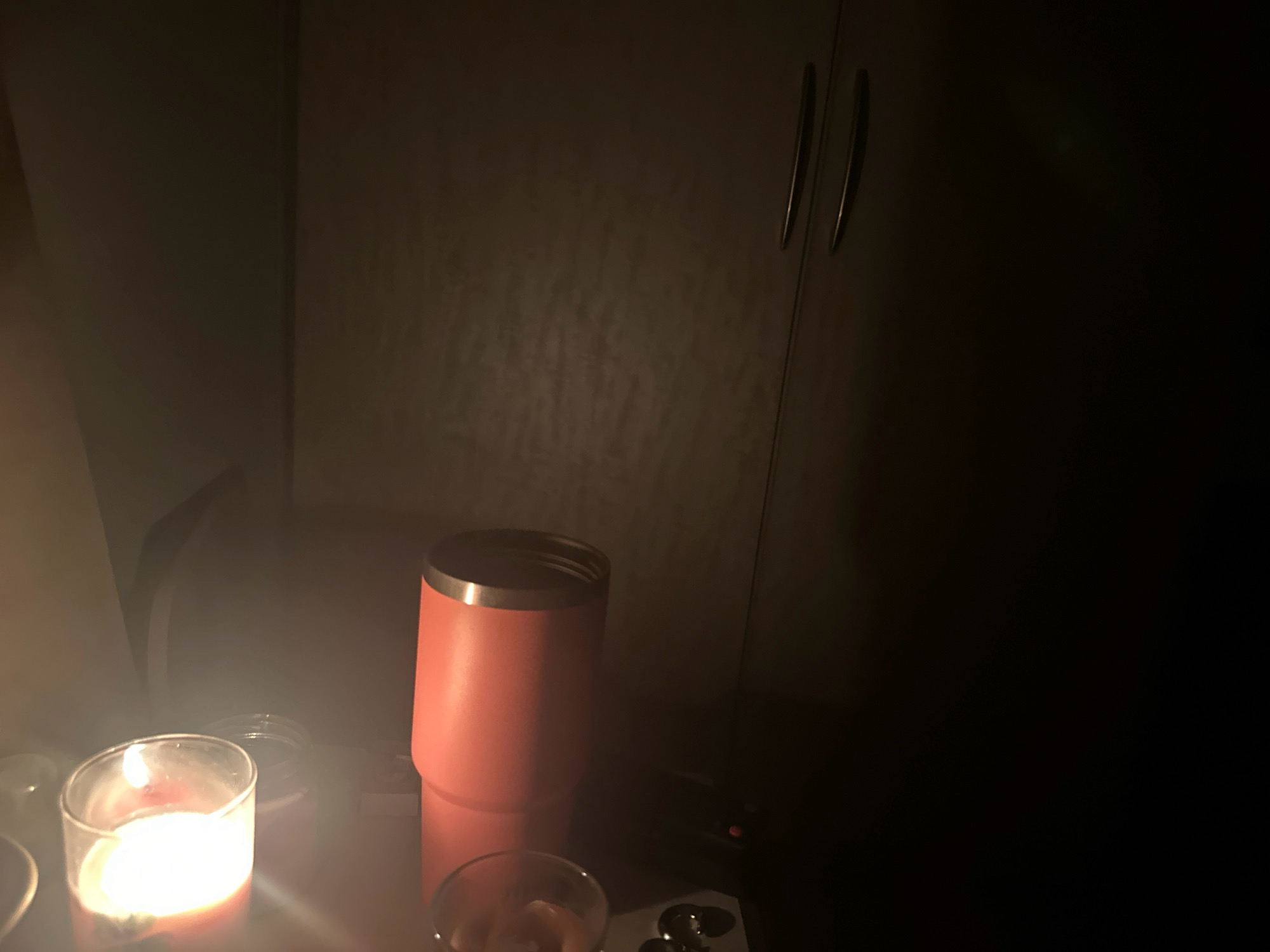 A lit candle in a glass container sits on a reflective surface in the foreground of a dark room. To the right of the candle is a tall, coral-colored tumbler with a silver rim. In the background, a dark wooden cabinet with two long, silver handles is visible. The overall lighting suggests the room is dimly lit, with the candle being the primary light source. There are no people present in the image. The location is not specified in the image, but if it were in Lalitpur, Nepal, it would depict an indoor scene.