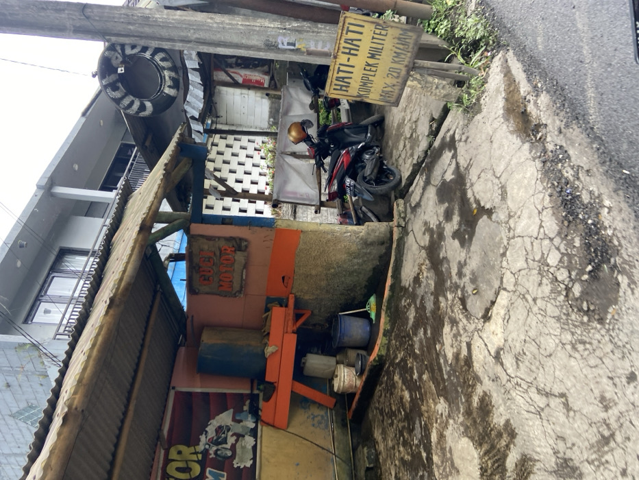 The scene shows a motorcycle parked outside a building in Lembang, Indonesia. The building has a sign that reads "Cuci Motor," indicating it is a motorcycle wash. The motorcycle is red and black, with a yellow helmet on the handlebars. There is a yellow sign with Indonesian text, likely a warning sign, next to the motorcycle. In the foreground, there is a muddy and cracked concrete area, likely a parking lot or driveway. There are also some buckets and other cleaning supplies visible. The image was captured at the coordinates -6.825 latitude and 107.61 longitude.