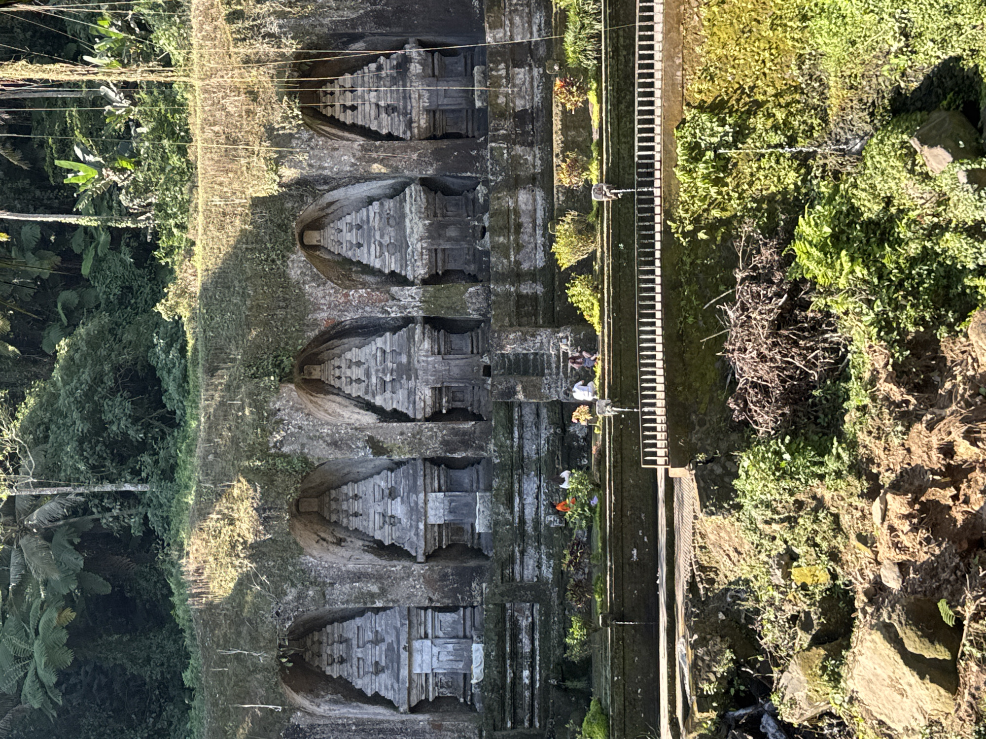 The image displays an ancient rock-cut temple complex set within a vibrant tropical landscape, indicative of locations like Tampaksiring, Indonesia. Five distinct shrine-like structures, each featuring a stepped pyramidal roof, are intricately carved directly into a vertical stone cliff face. These weathered carvings are partially covered with moss and other vegetation, blending with the natural environment. Stone platforms and steps lead up to and around these architectural elements.
To the right of the rock-cut temples, a sturdy walkway or bridge with railings extends horizontally, providing access along the site. The surrounding area is densely covered with lush green tropical foliage, including palm trees, ferns, and various broad-leafed plants that fill the background and foreground. A small body of water or stream is visible at the bottom right of the scene.
Several individuals are present at the site, appearing to be visitors or devotees. Two people are visible near the walkway, with one standing and another seemingly seated. Another person is closer to the temple carvings, possibly observing or participating in an activity. The scene is brightly illuminated by natural sunlight, suggesting either morning or late afternoon, which creates clear shadows and highlights. No discernible text is visible within the image.