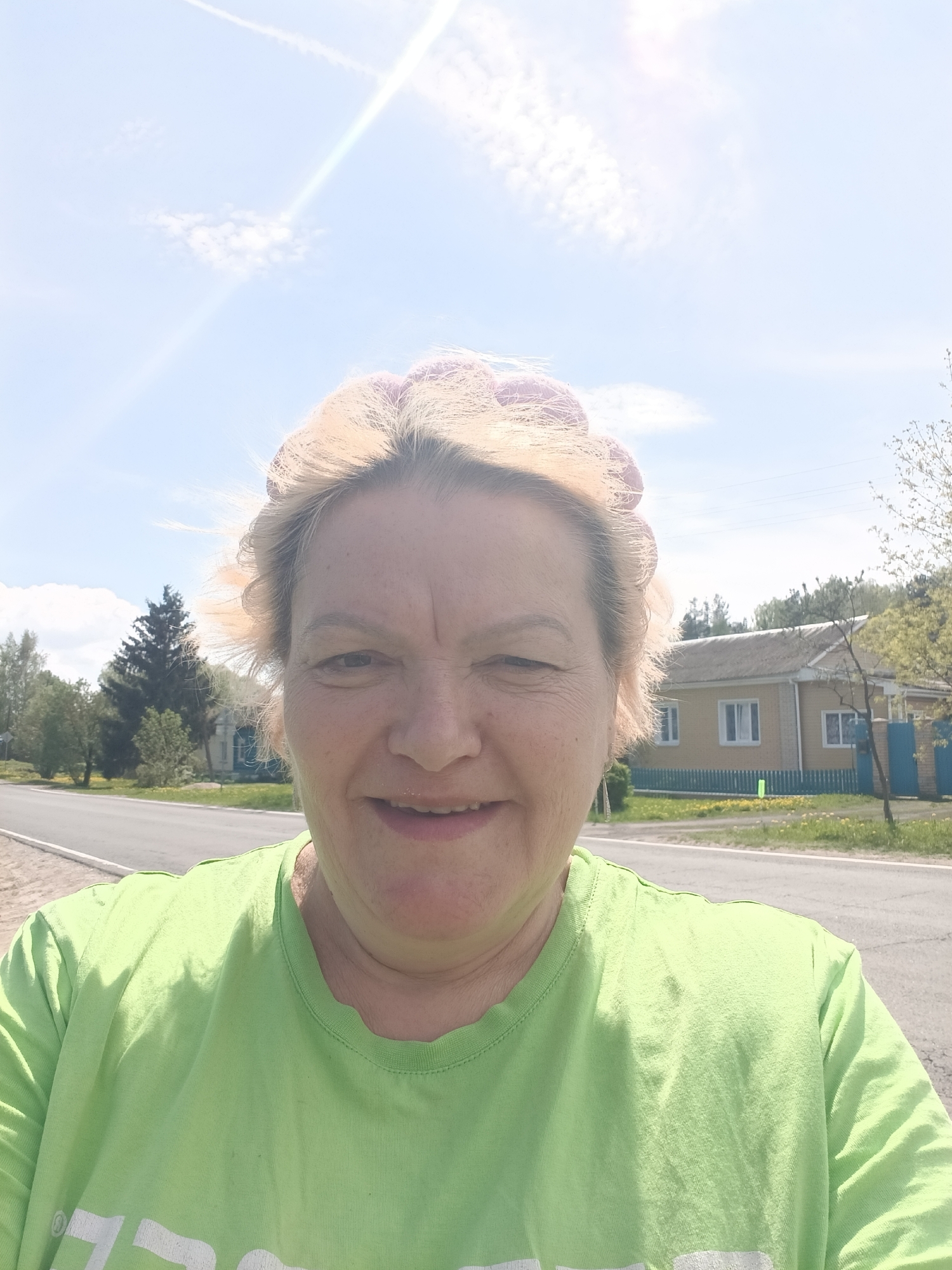 A woman is taking a selfie outdoors in Красный Маяк, Russia. She has light blonde hair with pink curlers. She is wearing a green T-shirt. A road runs behind her and a yellow house with a blue fence is visible. Trees are scattered in the background, under a bright sky.