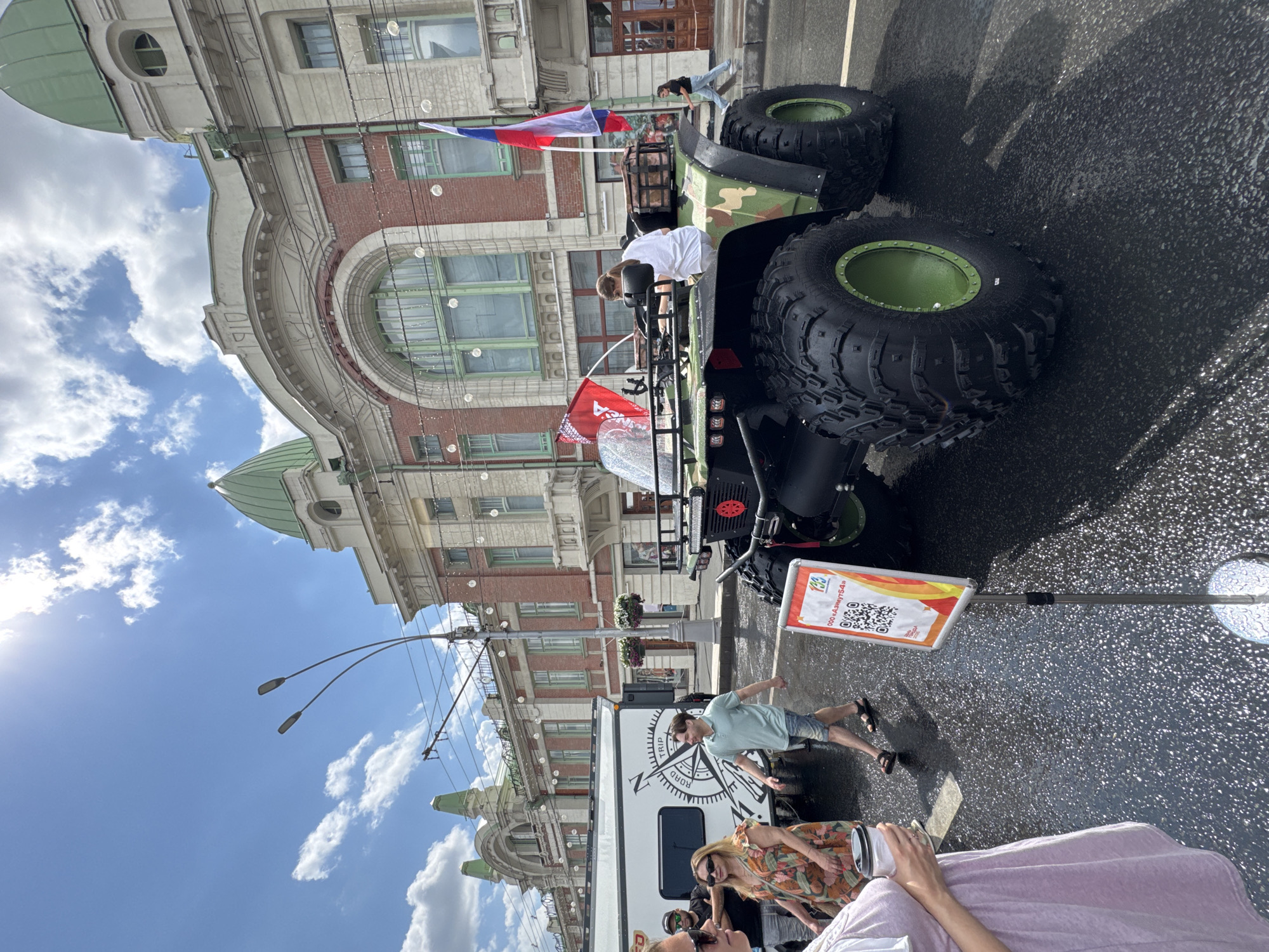 In a sunlit street scene in Novosibirsk, Russia, a large, camouflage-patterned all-terrain vehicle (ATV) with oversized tires is the central focus. The ATV, which appears to be a specialized expedition or off-road vehicle, is parked on a wet asphalt surface, indicating recent rain or street cleaning. Two Russian flags are prominently displayed on the vehicle, one a large tricolor and another smaller red flag, suggesting a patriotic or event-related display.
Several people are visible around the ATV and in the background. In the foreground on the left, a person in a light purple top holds a white cup. To their right, a woman in a floral dress and sunglasses stands near a white recreational vehicle (RV) adorned with a compass graphic labeled "ROAD TRIP". A man in a light blue polo shirt and denim shorts walks past them. Further in the background, a woman in a white shirt is seated on the ATV, and another person, possibly a man, is visible behind her. A few other individuals are also seen in the distance, including a person walking away from the ATV towards a building.
The backdrop consists of an ornate, historic building with red brick facade, arched windows with green frames, and decorative elements, characteristic of architectural styles found in cities like Novosibirsk. The building features green domes and what appears to be the year "1910" inscribed above a large archway, hinting at its age and historical significance. Streetlights and electrical wires crisscross the sky, and scattered white clouds are present in the bright blue sky. A sign with a QR code and Russian text is placed in front of the ATV, likely for promotional or informational purposes. The overall scene suggests an outdoor event or exhibition in a city center.
