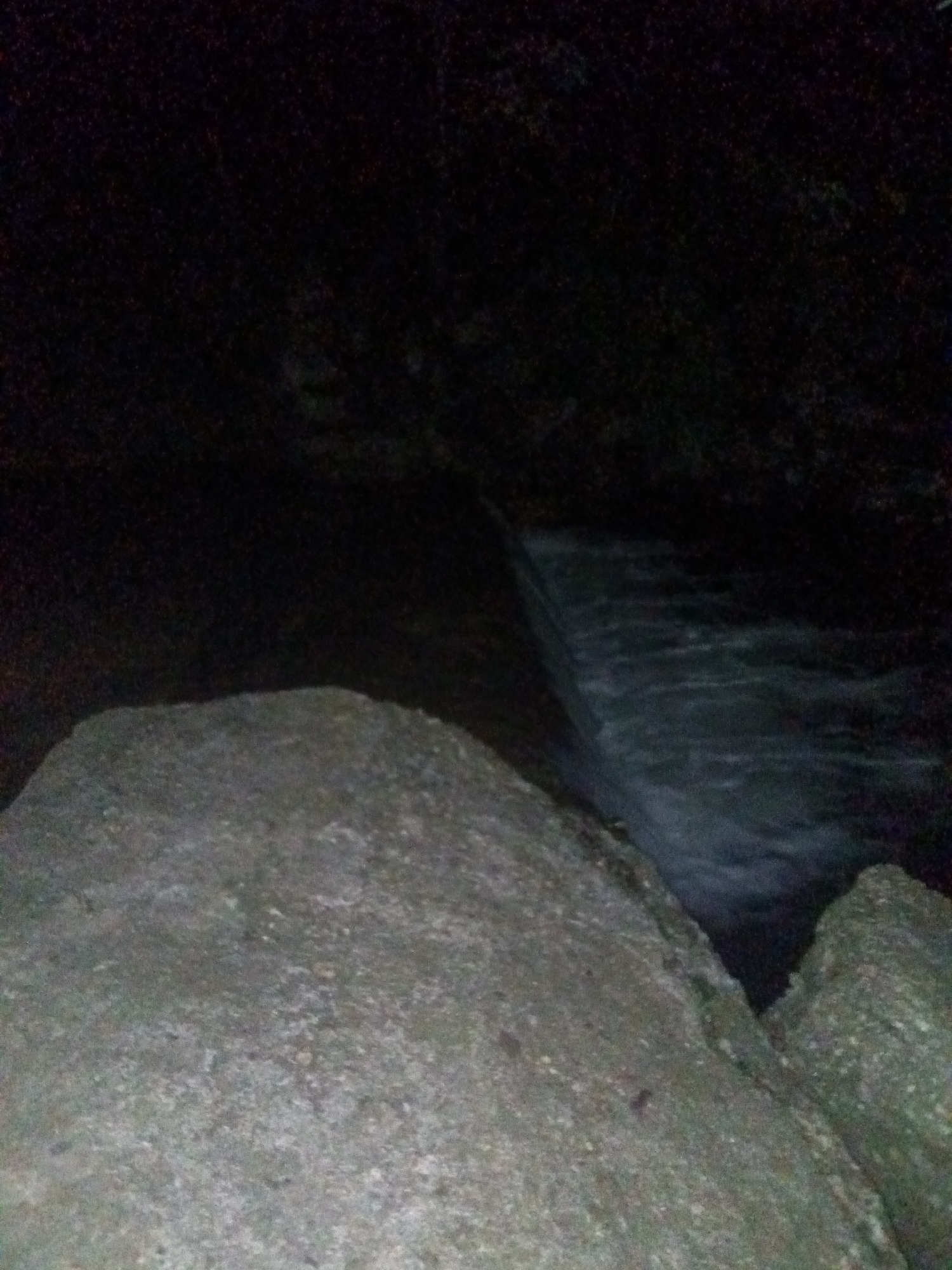 The image displays a dark, grainy night scene of a natural landscape, likely taken in Tahlequah, USA. The foreground is dominated by large, textured rocks or boulders, appearing in shades of grey and dark brown, occupying the lower half of the frame. These rocks show some rough texture and granular detail, hinting at a natural, rocky environment.
Behind the prominent rocks, a body of water, possibly a river or stream, flows. The water appears dark but shows signs of movement, with blurred ripples and slight reflections indicating its current, possibly forming a small waterfall or rapids as it descends slightly to the right side of the image.
The background is almost entirely enveloped in deep darkness, making it difficult to discern specific features. It appears to be an indistinct mass, possibly representing distant land, trees, or rock formations, all obscured by the lack of light.
The photograph was taken at night or very late dusk, as indicated by the extreme low light conditions and the overall blackness dominating the scene. There are no people, specific activities, or visible text in the image. The general atmosphere is one of quiet, dark natural beauty.