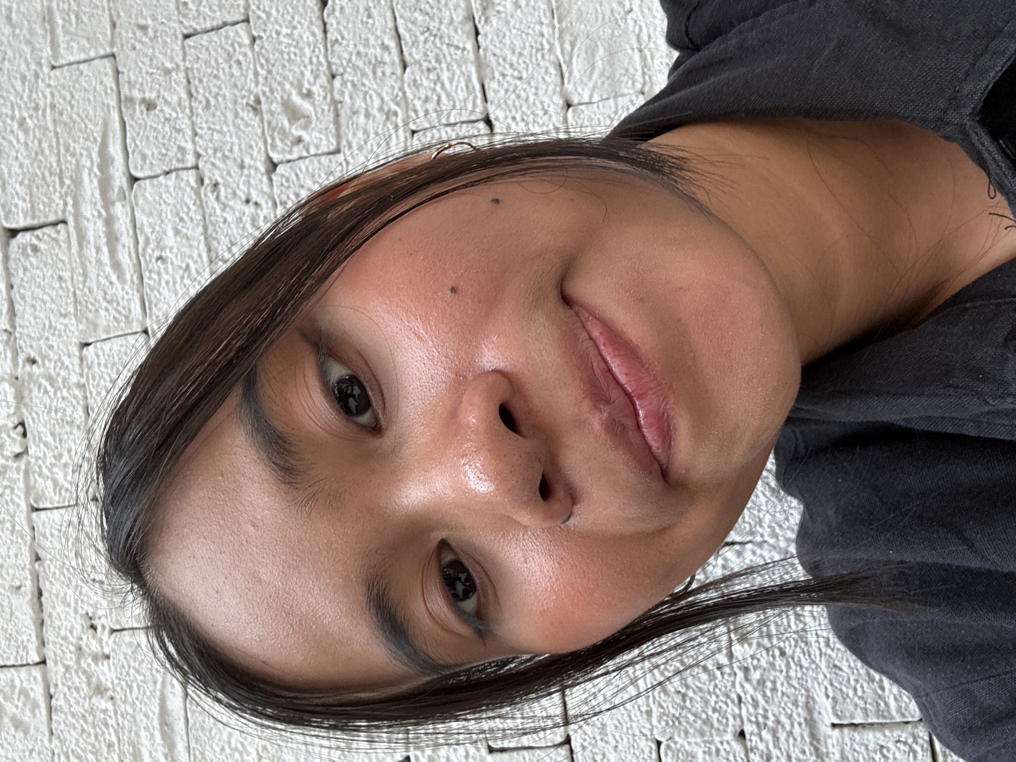 A close-up, high-angle selfie of a young woman with dark hair and brown eyes is shown. She is wearing a black collared shirt. Her face is tilted slightly, and she is looking towards the camera with a slight smile. Her skin appears to have some blush applied to her cheeks. She has two moles on her right cheek. The background is a textured white brick wall. The lighting suggests an outdoor setting, possibly during the day. The location is South Jakarta, Indonesia.