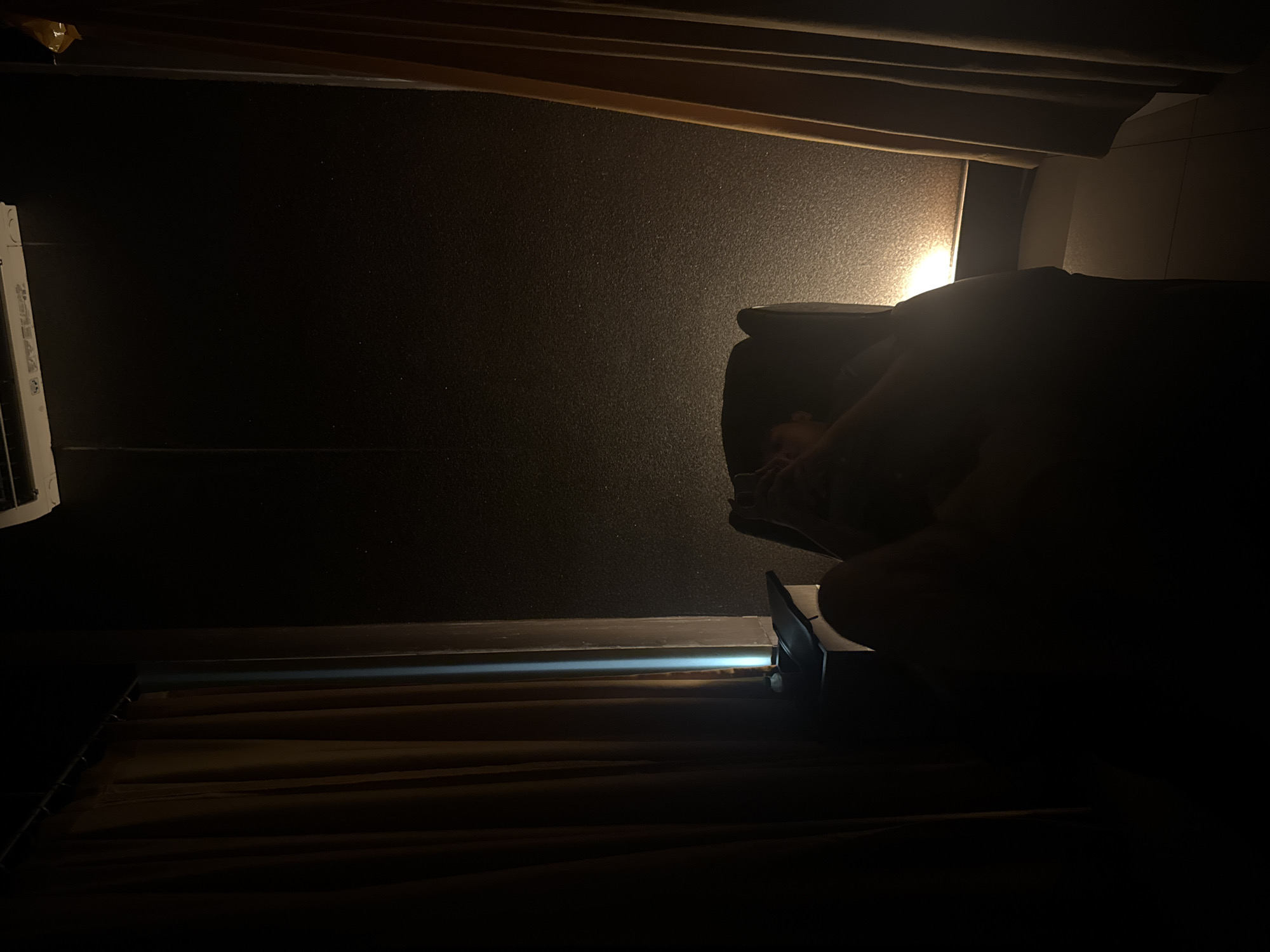 The image shows a dimly lit interior space. A person is seated in what appears to be a chair, though only the upper portion of their body is visible and largely obscured by shadow. The background is a dark wall, possibly textured. Below the wall is a horizontal surface, lit by a faint light source, and covered with what appear to be folded fabrics. The location is Gunung Putri, Indonesia.