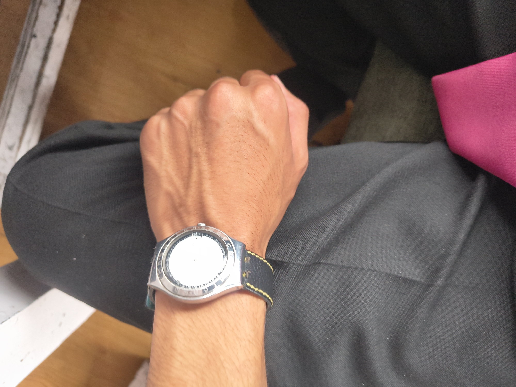 A close-up, high-angle shot shows a person's wrist wearing a silver-toned watch with a black band and yellow stitching. The watch face is white with black markings. The person is wearing black trousers. The background consists of a wooden floor and a white surface that appears to be part of a bench or seating area. A bright pink object is visible in the lower right corner. The image is taken in an indoor setting. No specific location in Dharamshala, India can be determined.