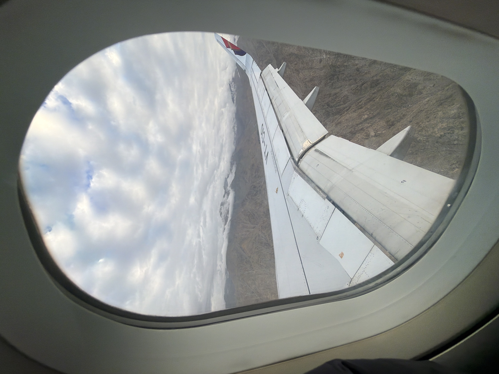 This is an aerial view of the city of Leh, India, taken from an airplane window. The image features a prominent airplane wing in the foreground, extending across the frame. Beyond the wing, the rugged, mountainous landscape of Leh is visible, with some areas appearing barren and rocky. Clouds are present in the sky, partially obscuring the distant mountains. The overall scene depicts a flight over the mountainous terrain surrounding Leh.