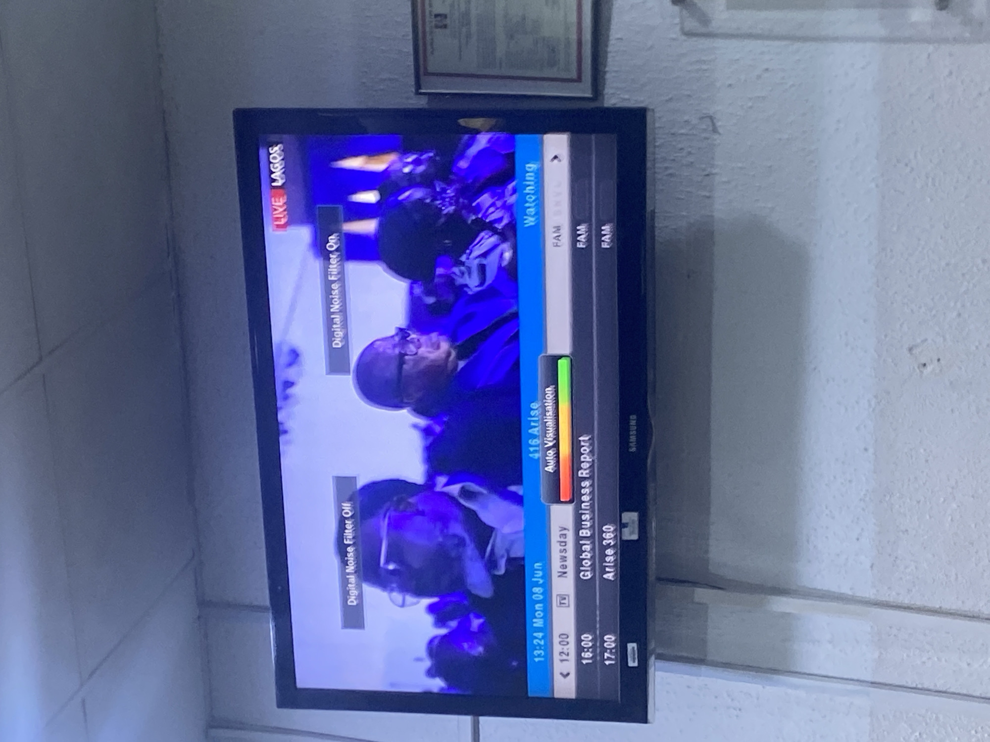 A television screen displays a news broadcast with a "LIVE LAGOS" banner. The screen shows a program guide with times, channels, and titles such as "Newsday," "Global Business Report," and "Arise 360." The television is mounted on a white, textured wall.