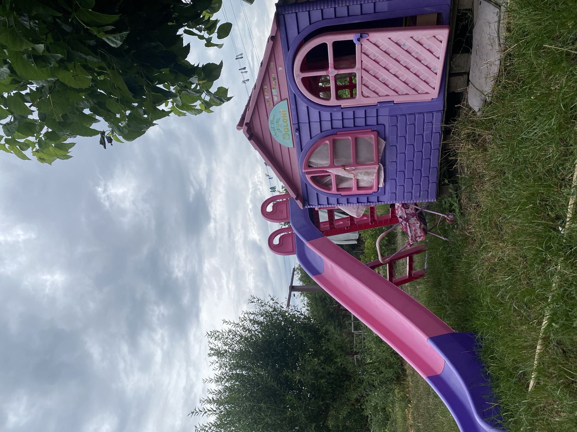 The image shows a purple and pink children's playhouse with a slide in a grassy backyard. The playhouse has two arched windows and a door with a diamond pattern. Above the windows, there is a sign that says "Sweet Home BOLONI" with some butterfly decals. A pink slide is attached to the side of the playhouse, with purple railings. In the background, there is a large green bush, some trees, and a cloudy sky. There is also a clothesline with several items of clothing hanging on it. The scene appears to be in a residential area, likely a backyard in Kropyvnytskyi, Ukraine, given the provided context. There are no people visible in the image. The overall impression is of a slightly overcast day in a typical suburban or rural setting.