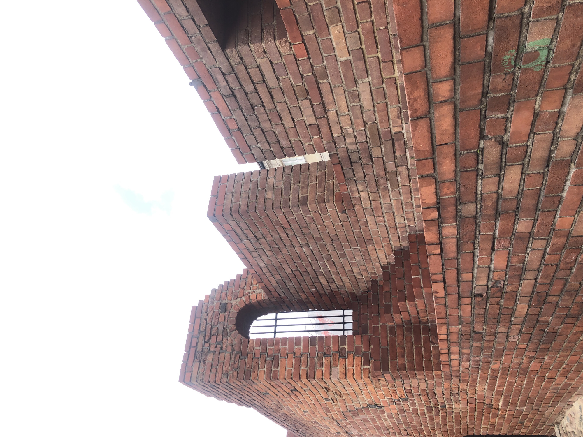 The image depicts a section of a red brick wall, possibly part of a historic fortification or building, located in Warsaw, Poland. The wall is constructed with weathered red bricks, showing variations in color and texture. In the foreground, the wall extends diagonally from the bottom left to the top right of the frame, with a large expanse of bricks. There is a stepped indentation in the wall on the left side, leading up to an arched opening. This opening is fitted with a dark metal grate, behind which some indistinct shapes and colors, possibly a sign or advertisement, are visible. The sky above is overcast and bright, with a hint of blue and clouds visible in the upper right portion of the image. The overall impression is of an old, possibly renovated, defensive structure.
