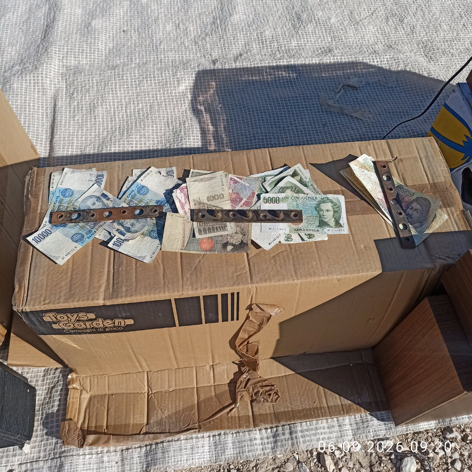 In the town of Sannicandro di Bari, Italy, an outdoor scene depicts a cardboard box opened and placed on a textured white surface. The box contains a collection of various currency notes, including Italian Lire. A metal strip with six circular holes is placed horizontally across some of the banknotes. The environment appears to be a market or outdoor area, with shadows cast by an unseen object. The date "06.09.2026 09:20" is superimposed on the lower right of the image. The box is labeled "Toys Garden Compagni di gioco."
