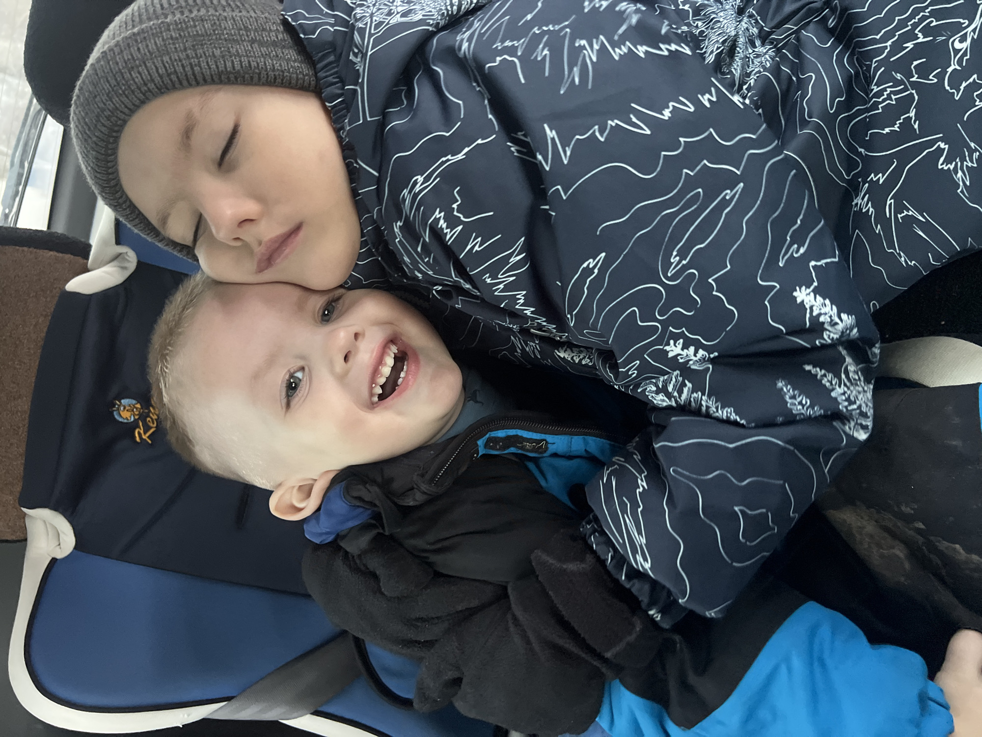 The image captures two children inside a vehicle. The precise location within the city of Chaikovsky, Russia, cannot be determined from the visual evidence.
A younger child, with light hair and blue eyes, is positioned on the left, looking directly at the camera with an open-mouthed smile, revealing upper teeth. He wears a black puffer jacket over a blue undergarment and black gloves. He is partially seated in a blue and dark blue child booster seat, secured by a grey seatbelt. A yellow logo depicting an animal and the text "Ken" is visible on the booster seat's headrest.
An older child is positioned on the right, with closed eyes, resting his head against the younger child's head. He wears a grey knitted beanie and a dark blue jacket featuring a white line-art pattern of mountains and foliage. The children appear to be in close physical contact, suggesting an embrace or a shared moment of rest. The background shows elements consistent with a vehicle interior, including a window line.
