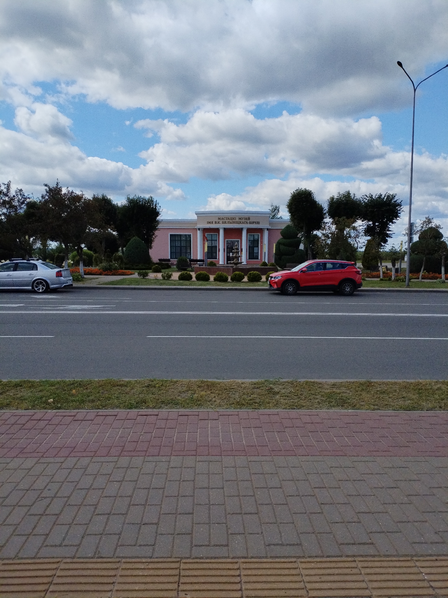 The image depicts a street scene in Bialynichy, Belarus, featuring a light pink building with columns and text identifying it as the "MASTACKI MUSEUM IMIA V.K. BYALINICKAGA-BIRULI." A silver sedan is parked to the left of the building, and a red SUV is parked in front of it, to the right. The street in the foreground has multiple lanes, bordered by a grassy median and a paved walkway. Trees and manicured bushes surround the museum, and a lamppost stands tall on the right side of the image. The sky is partially covered with white and gray clouds.