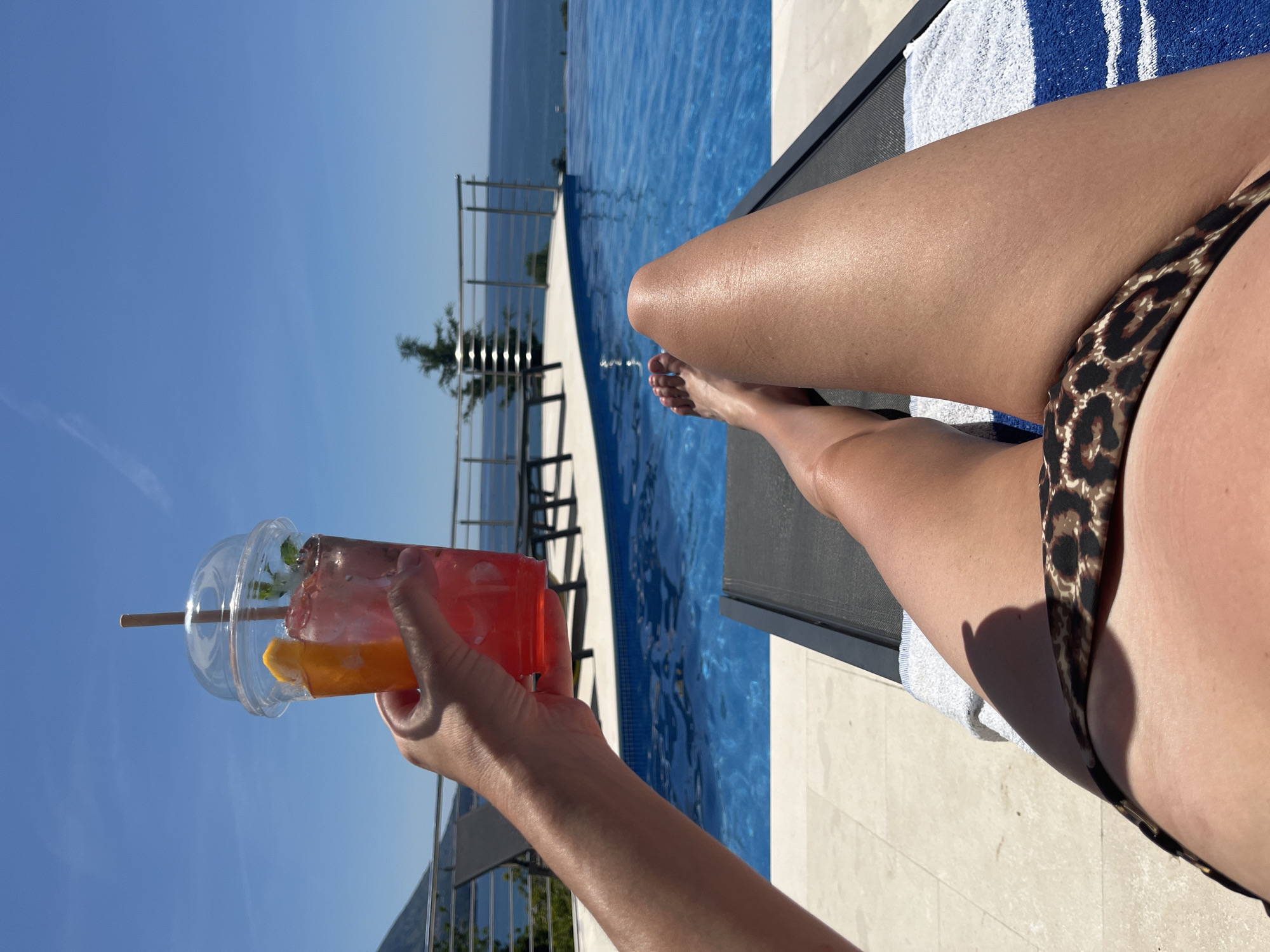 A person is lying on a lounge chair, possibly sunbathing, next to a pool in Boreti, Montenegro. The person is wearing a bikini bottom with an animal print. The pool is blue and has a view of the sea. The person is holding a clear plastic cup with a red drink, ice, an orange slice, and a straw. The sky is blue and clear. A metal railing and a tree are visible in the background. A white and blue striped towel is on the lounge chair.