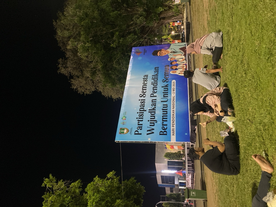 The image shows a group of four people sitting on a grassy area in front of a large billboard at night. The billboard is blue and white, with the text "Partisipasi Semesta Wujudkan Pendidikan Bermutu Untuk Semua" in large, bold letters. Below the text is the smaller text "HARI PENDIDIKAN NASIONAL - 2 MEI 2023." The billboard features a group of smiling people, some of whom are wearing traditional Indonesian clothing. The four individuals seated on the grass are all wearing casual clothing, and two of them are looking at the billboard, while two others are looking at each other. The location is Surakarta, Indonesia, at the coordinates -7.569 latitude and 110.83 longitude. The background consists of a dark sky, lush foliage, and a nearby building.